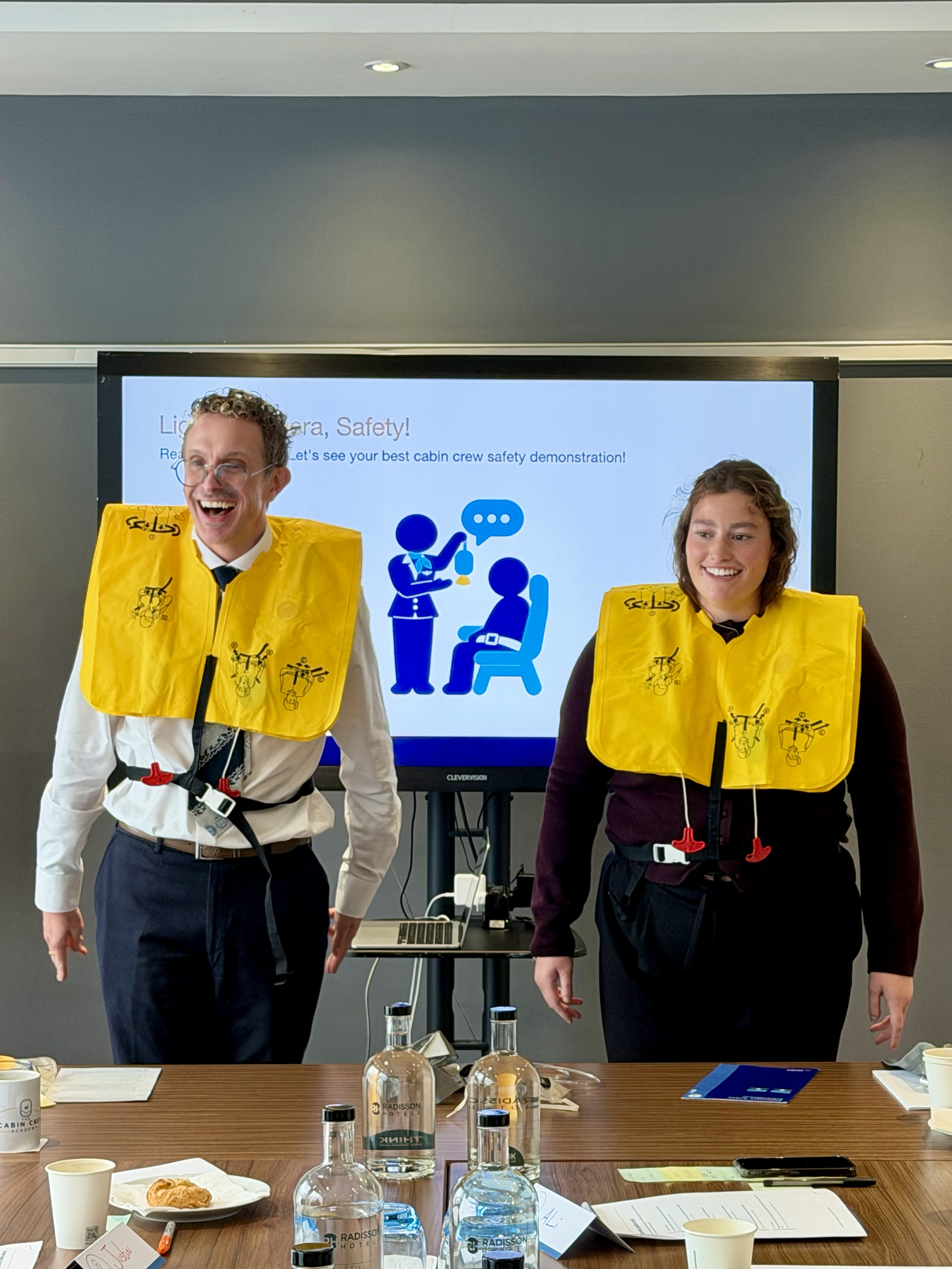 Two cabin crew trainers wearing yellow airline safety vests stand in front of a cabin safety demonstration presentation.