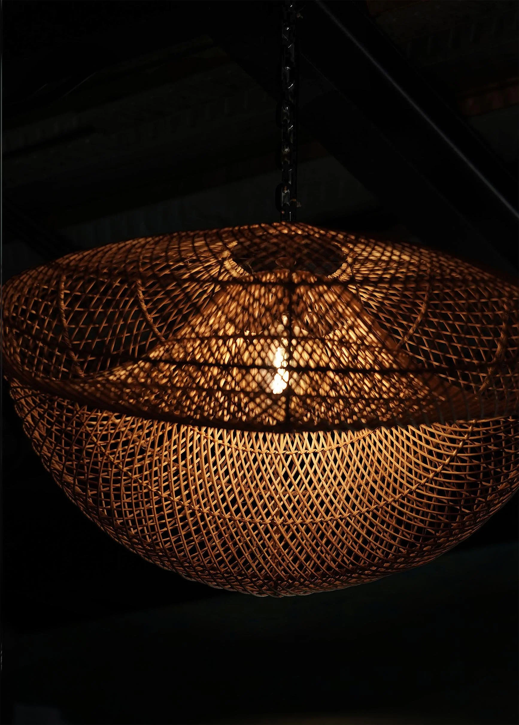 A woven rattan pendant light with a warm glow hangs from a chain in a dark setting.