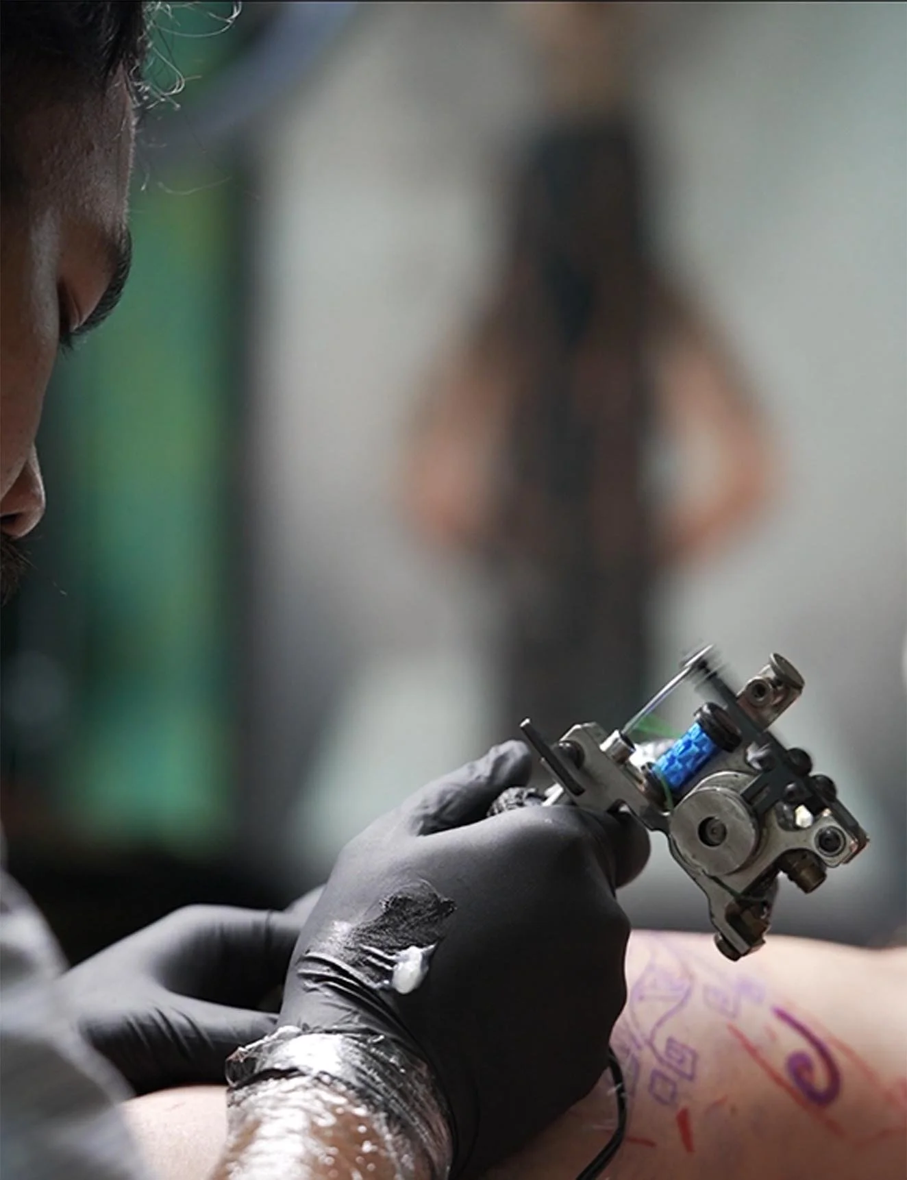 Close-up of a person wearing black gloves, using a tattoo machine with a needle, on someone's skin. The person is focused, and there are colorful tattoo designs on the skin.