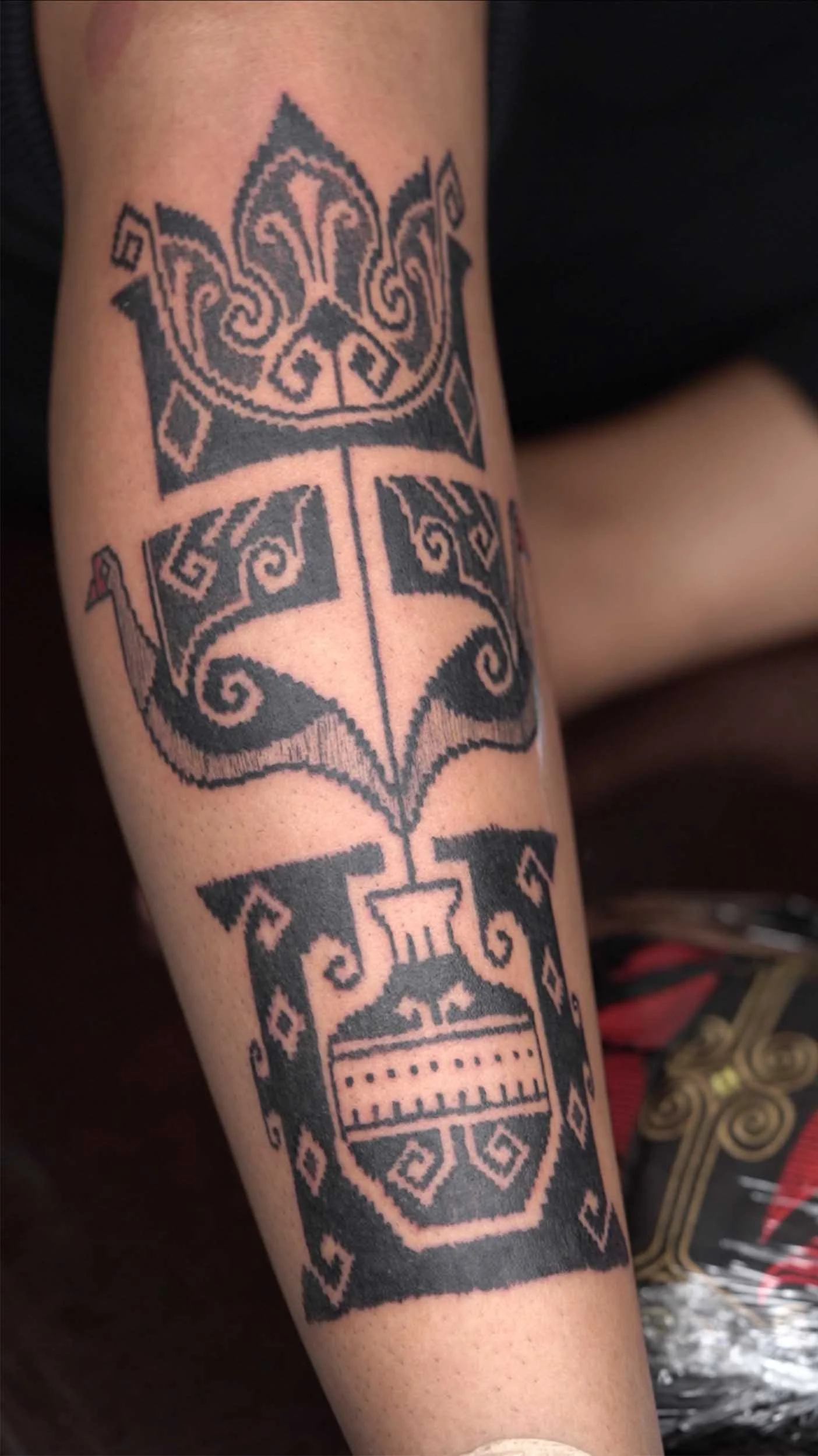 A black tribal-style tattoo on a person's shin featuring a symmetrical design with sharp angles, curved lines, and intricate patterns