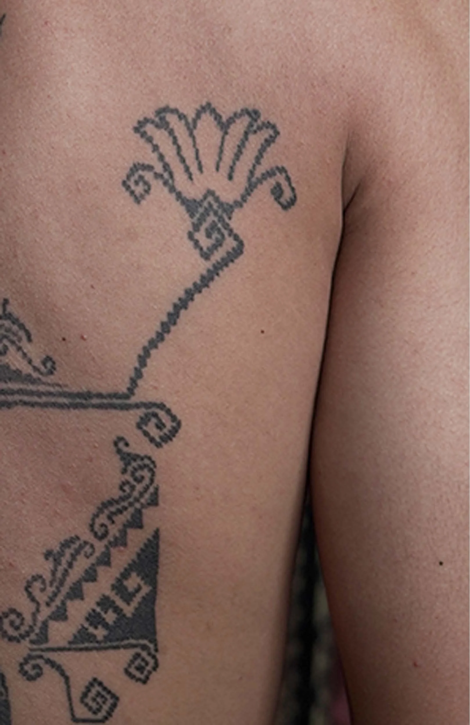 Close-up of a person's upper chest and shoulder showing a black tattoo of a stylized flower with decorative elements.