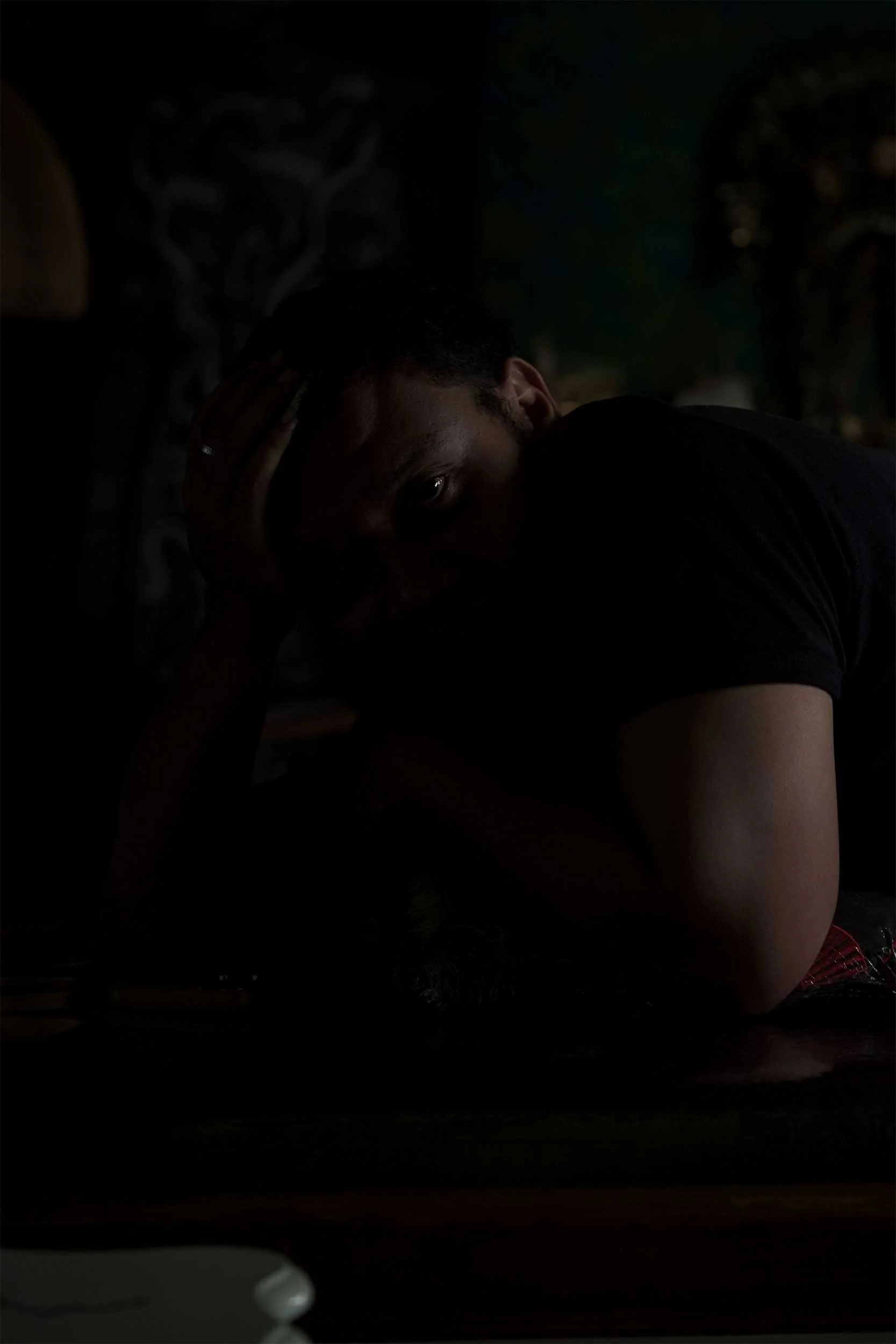 A person with short hair and a dark shirt resting their head on a surface, looking directly at the camera in a dimly lit room.