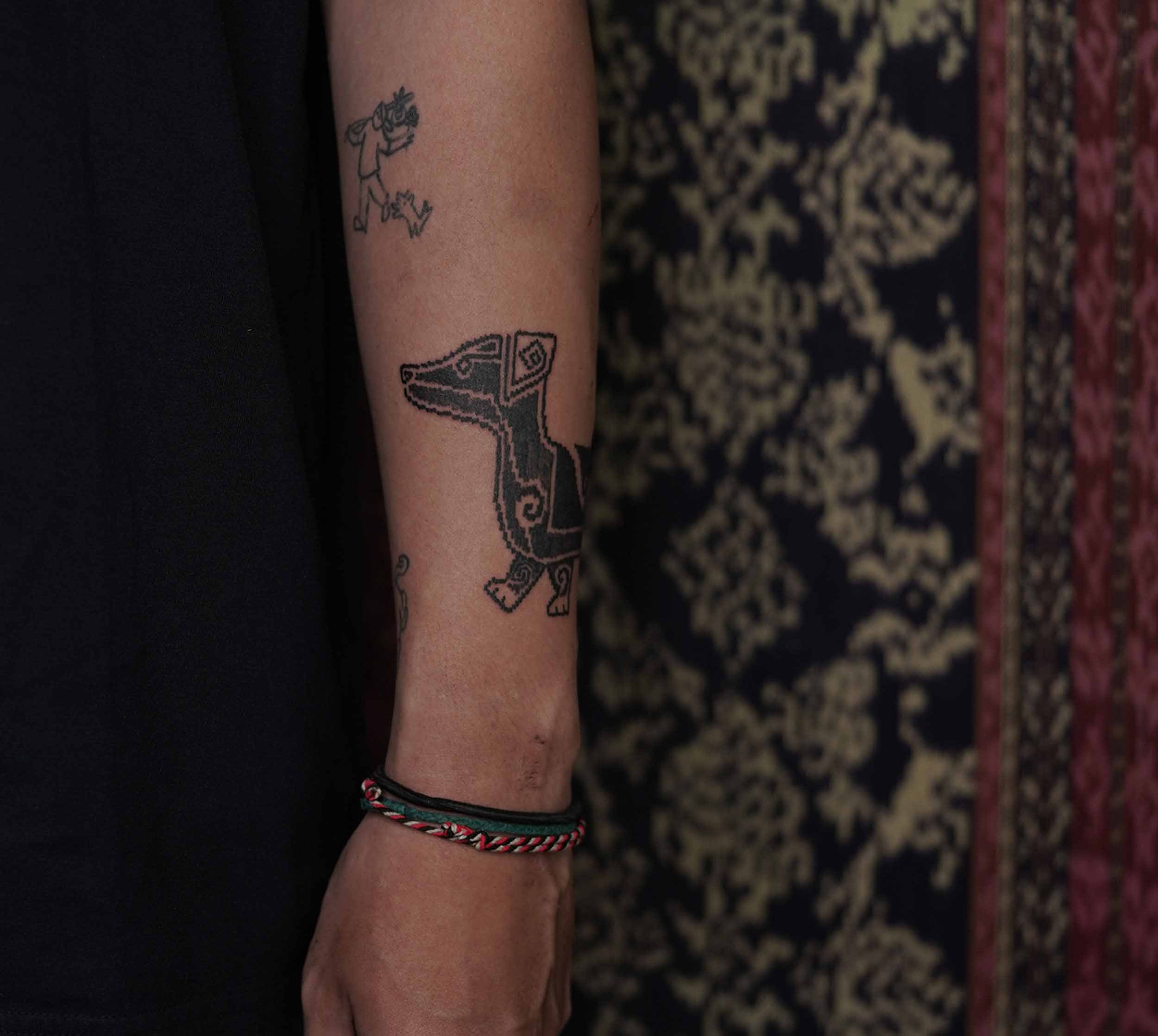 A person's forearm with a tattoo of a cartoon-style whale and a small figure of a girl with a rose in her hair. The person is wearing a black shirt and a red, black, and white woven bracelet on their wrist. The background shows a patterned carpet.