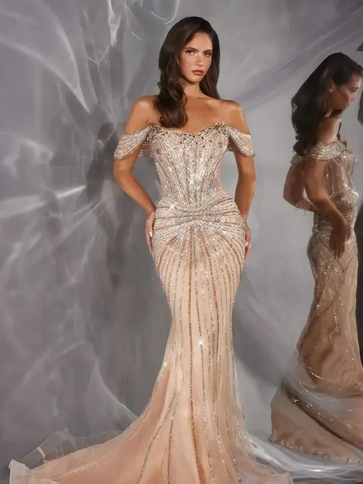 A woman in a glamorous, strapless, sparkly evening gown with a mermaid silhouette, standing against a gray background with her reflection visible in a mirror.