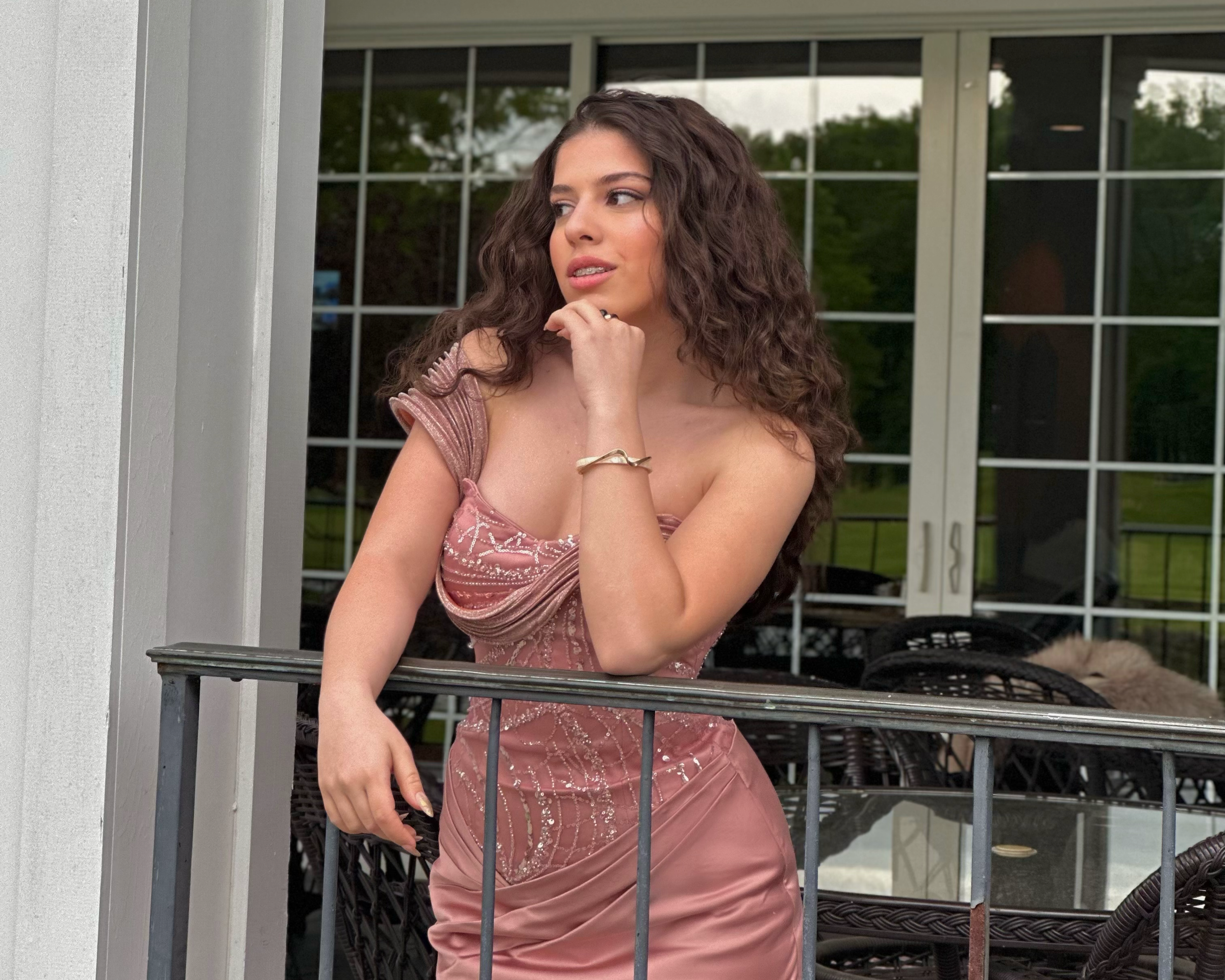 A young woman with curly brown hair standing behind a balcony railing on an outdoor patio. She is wearing a pink dress with lace details and jewelry including rings and a bracelet. In the background, there is a large window with trees visible outside.