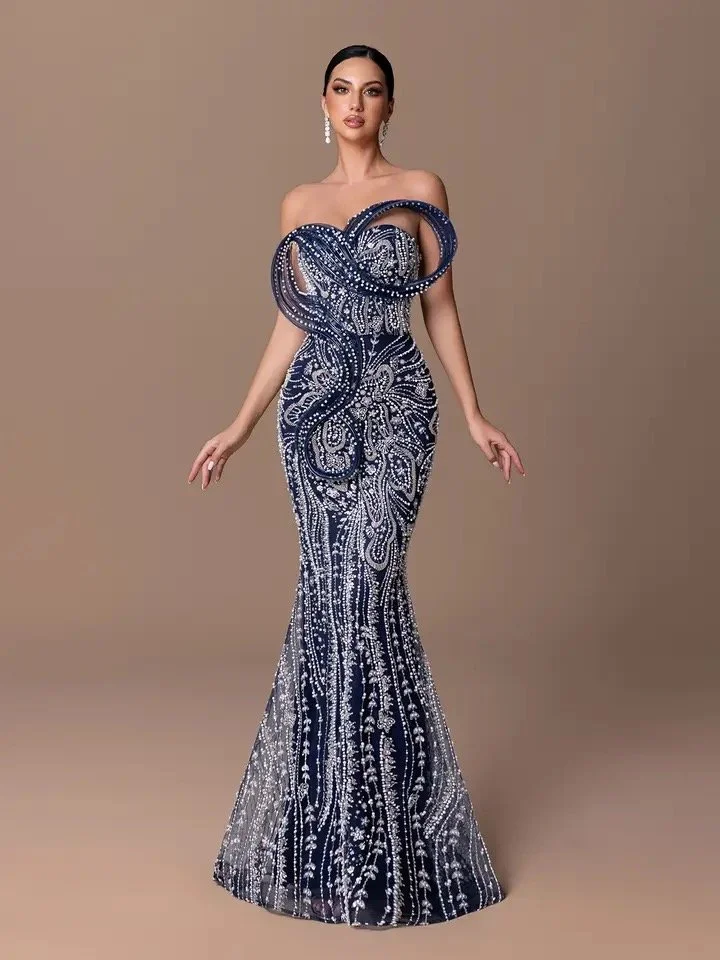 A woman wearing an elegant, off-the-shoulder navy and silver beaded evening gown with intricate patterns and large decorative loops on the chest, standing against a plain background.