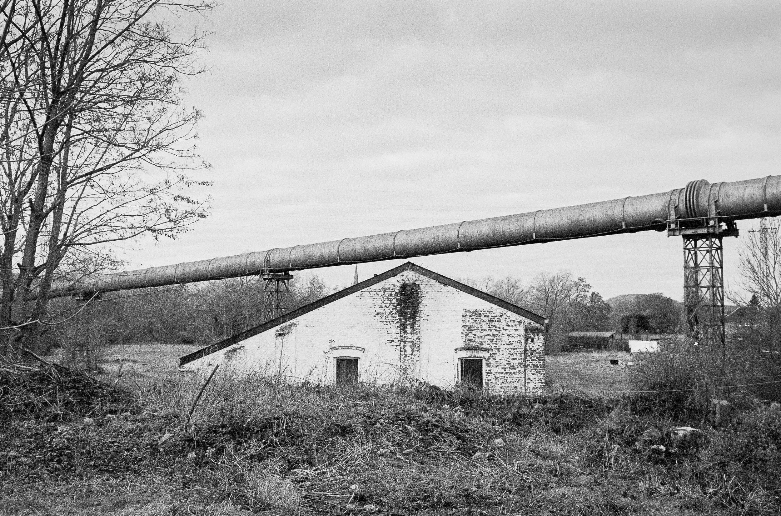 Black and white photo of a rural scene with a brick house, leafless trees, and a large pipe running horizontally across the scene above the house.