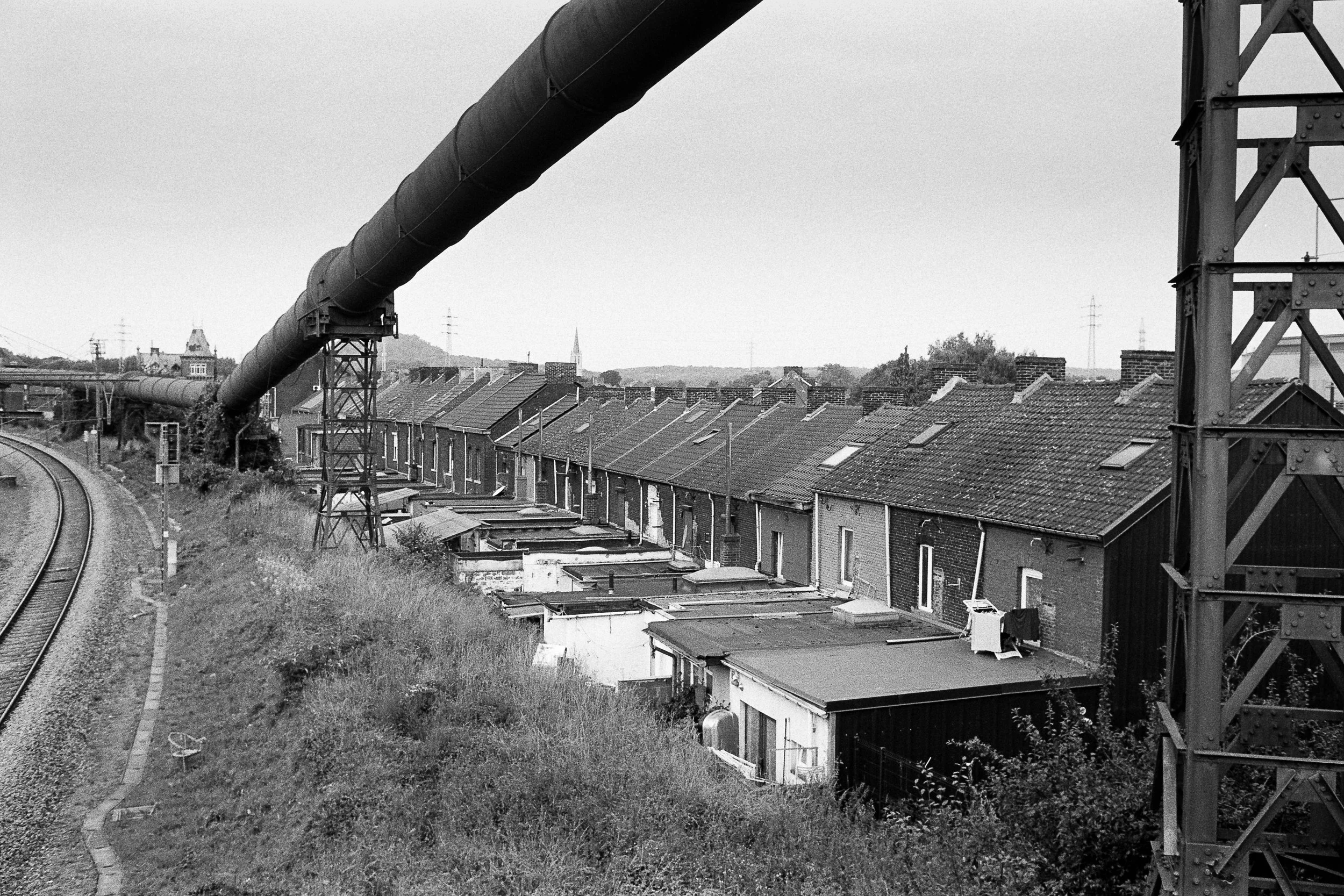Black and white photo of a row of houses with a large industrial pipe running above them and a metal structure supporting the pipe. The background shows trees and power lines.