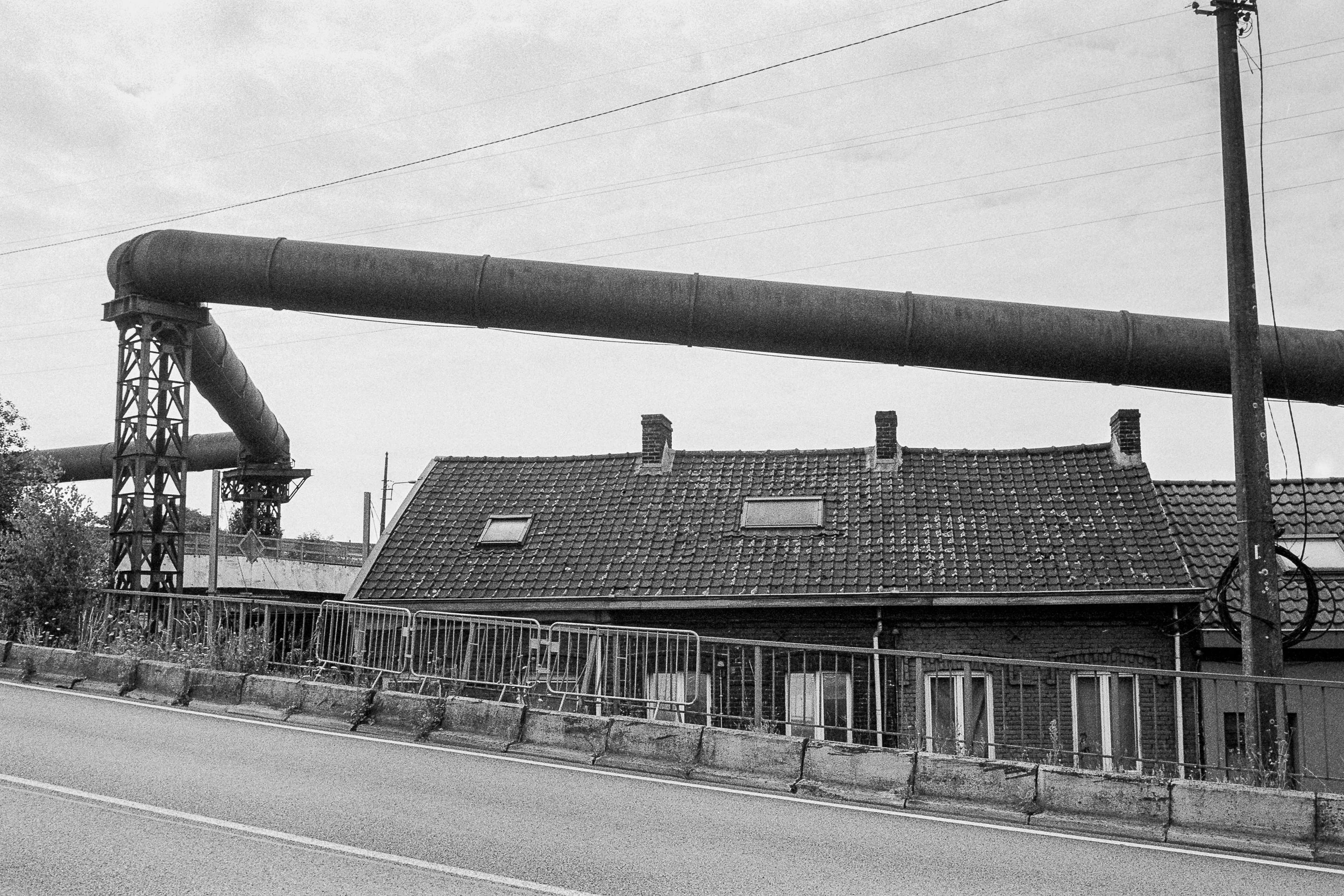 A black and white photo of a large industrial pipe running horizontally above rooftops, with a utility pole and power lines in the background.