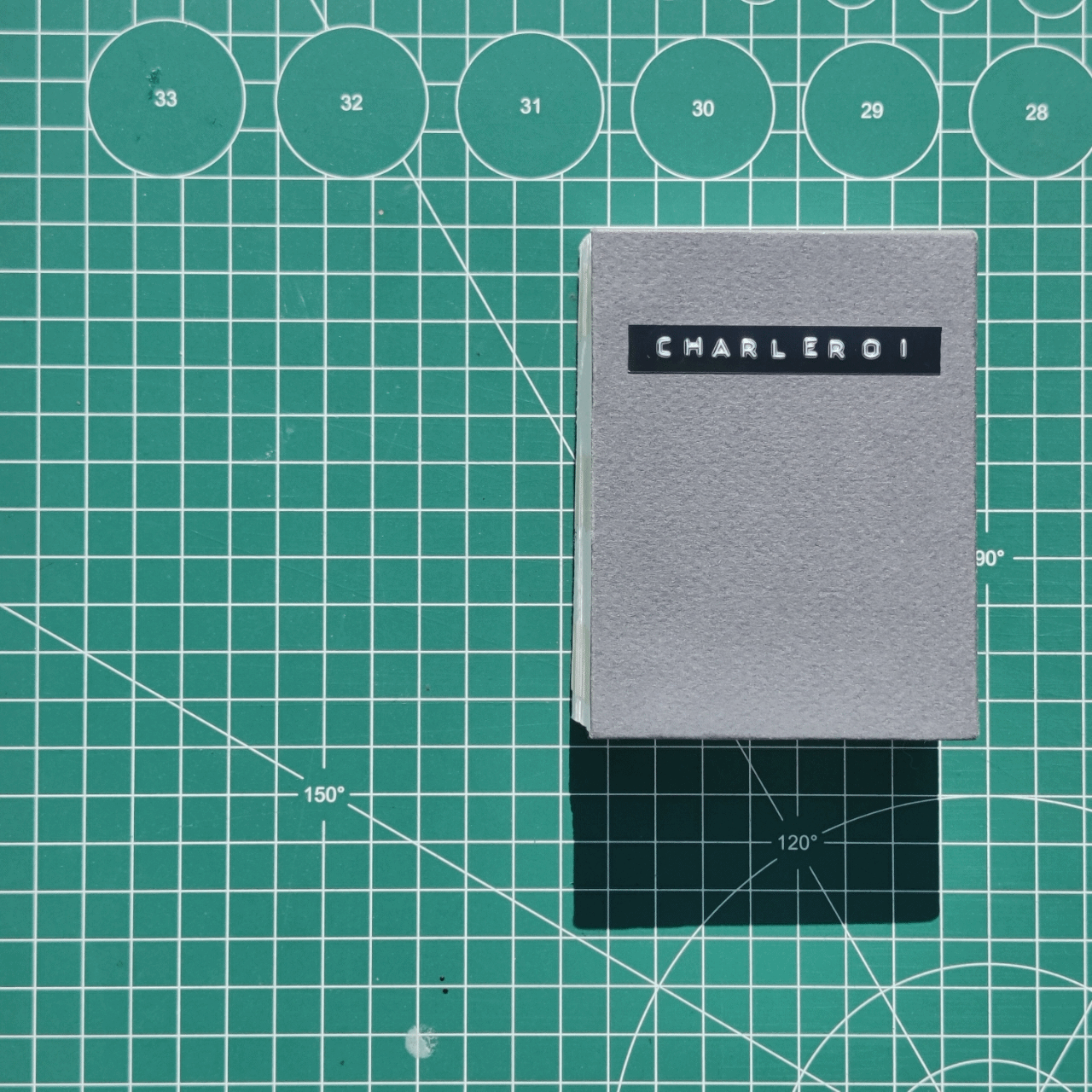 A gray book is placed on a green cutting mat with a grid and circle measurements. The book has 'CHARLEROI' written on a black label on its cover, and its shadow is cast on the mat.