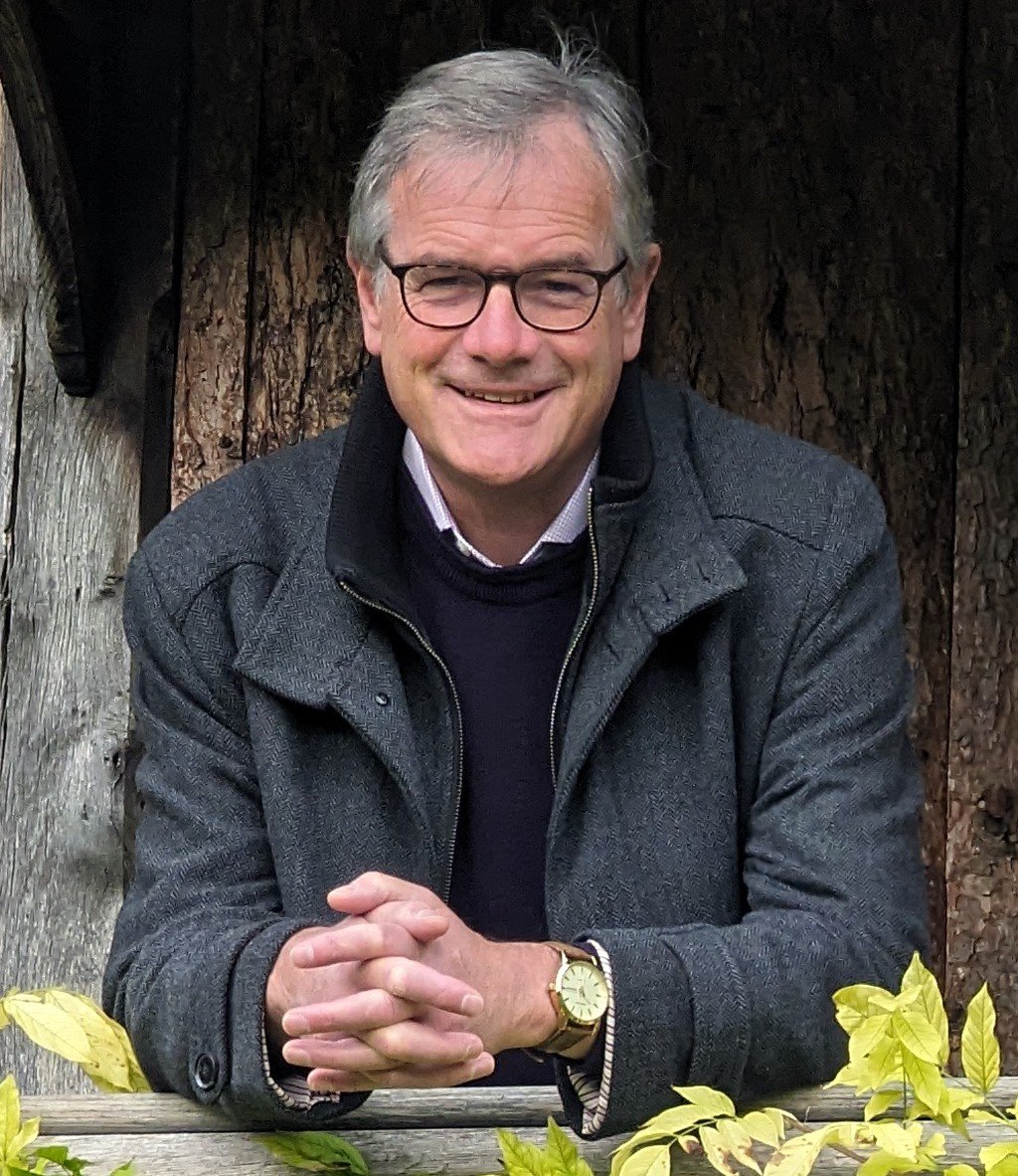 A smiling middle-aged man with glasses and gray hair, wearing a dark gray coat and a gold watch, sitting outdoors at a wooden table in front of a large tree trunk and yellow leaves.
