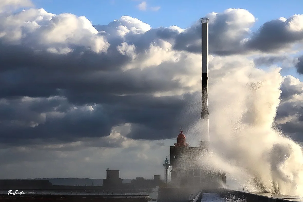 Photo magnifique — phare + vague en brume + ciel dramatique bleu ! 
`La vague s'écrase. Le phare disparaît dans la brume blanche. Et pourtant, il tient.`