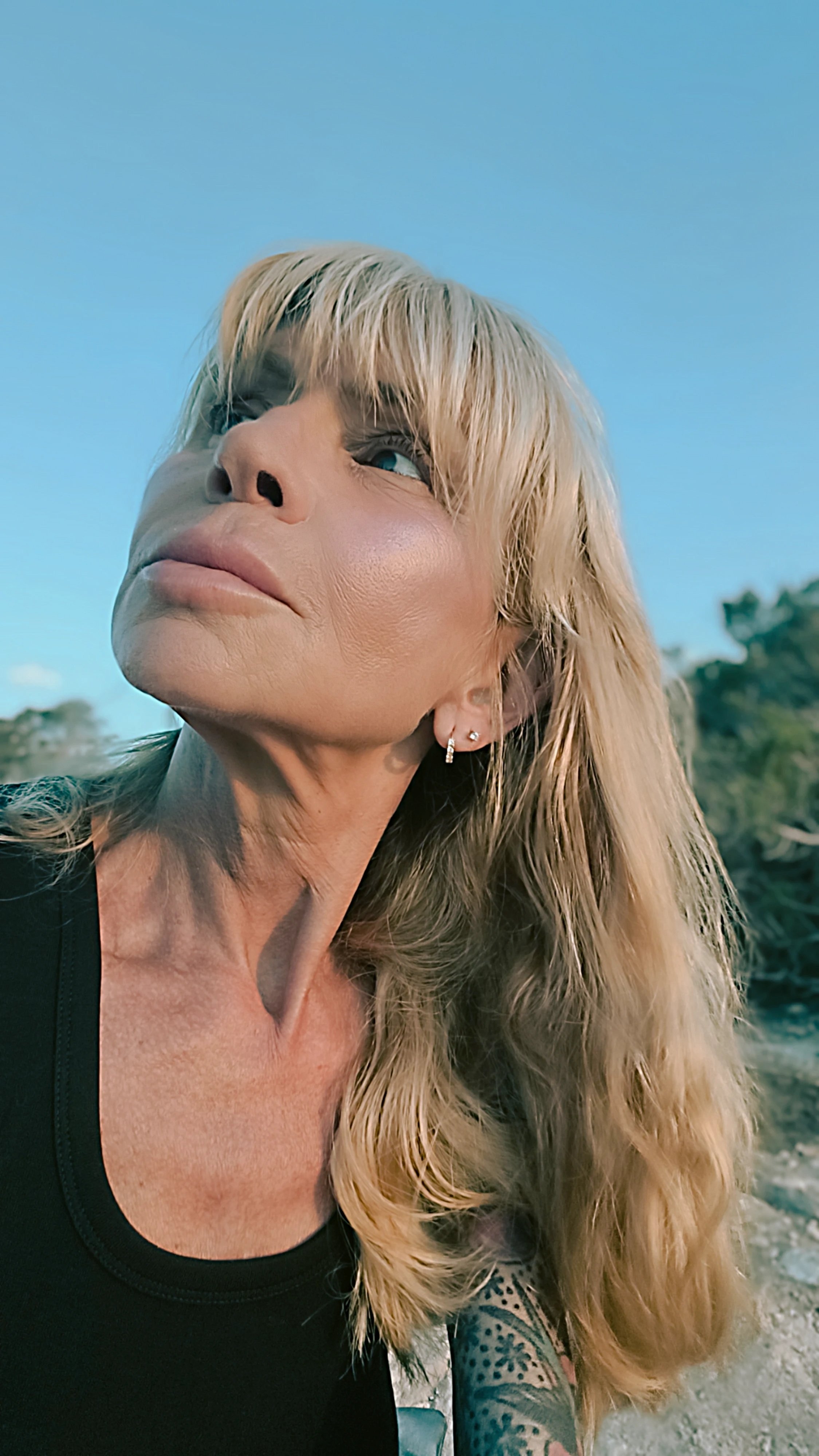 A woman with blonde hair and tattoos on her right arm, wearing earrings and a black top, outdoors with clear blue sky and trees in the background.