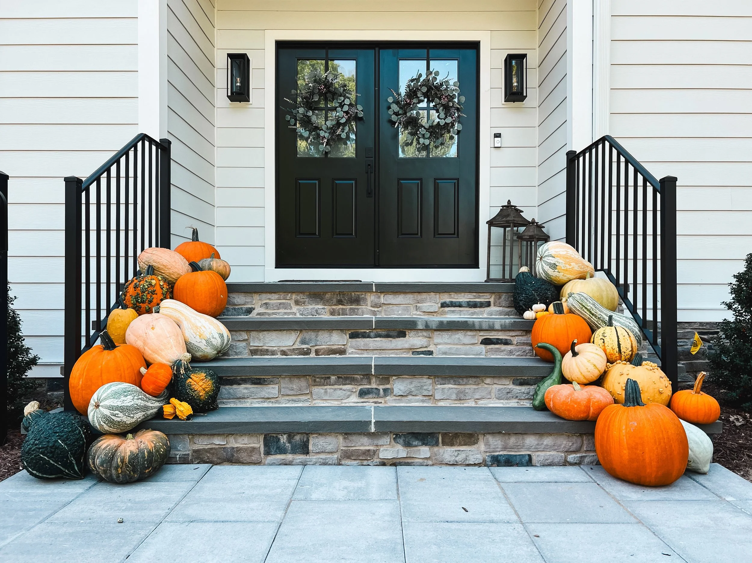 Front porch with black double doors decorated with wreaths, surrounded by white siding, black railings, and an assortment of pumpkins and gourds on the steps and ground in front.