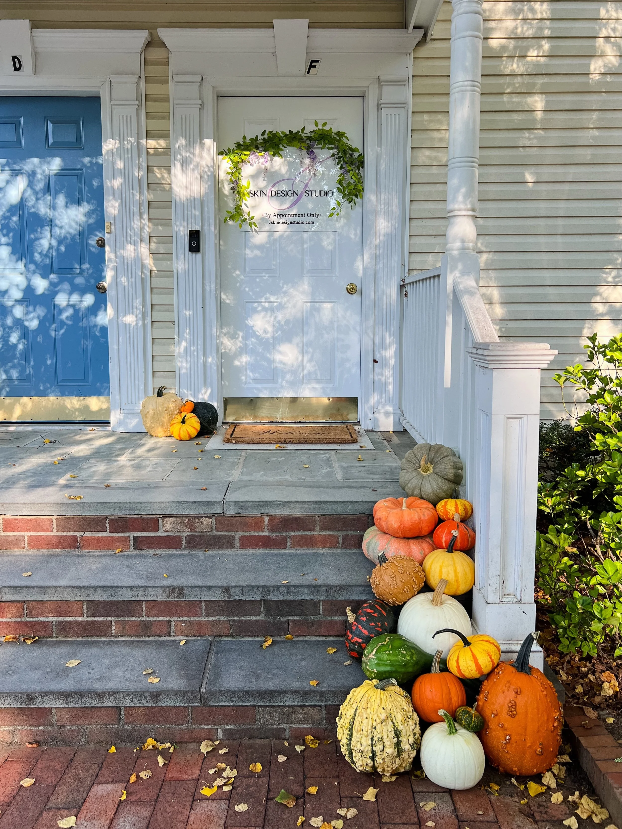 Front porch of a house with a white door and a blue door beside it. Fall pumpkins of various sizes and colors are arranged on the steps and ground near the porch railing, with fallen leaves scattered around.