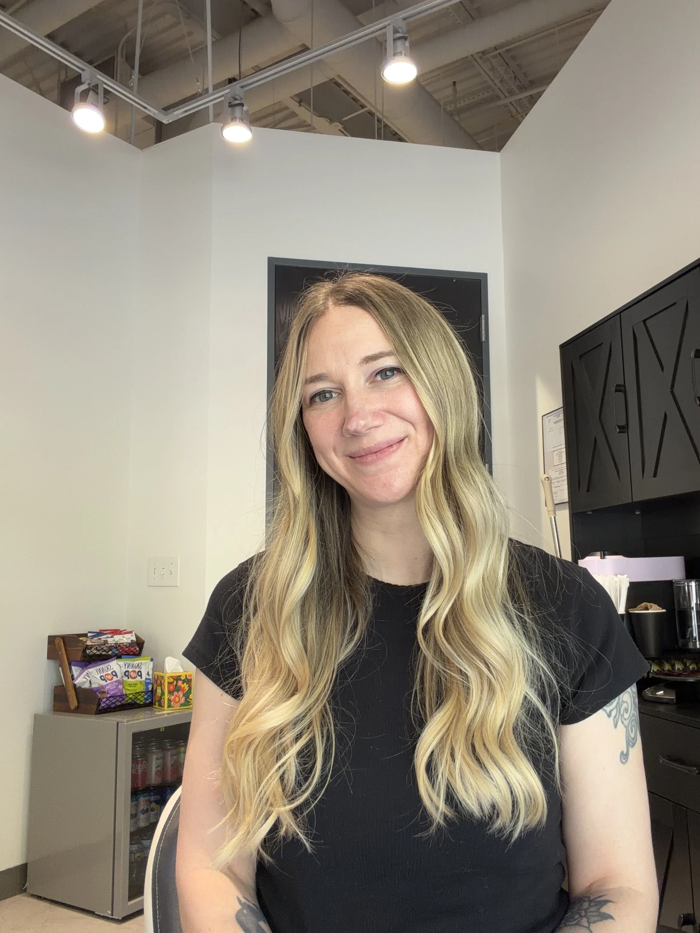 A woman with long blond hair sitting in an office or break room, smiling at the camera. Behind her are shelves stocked with snacks and drinks, and a black cabinet to her right.