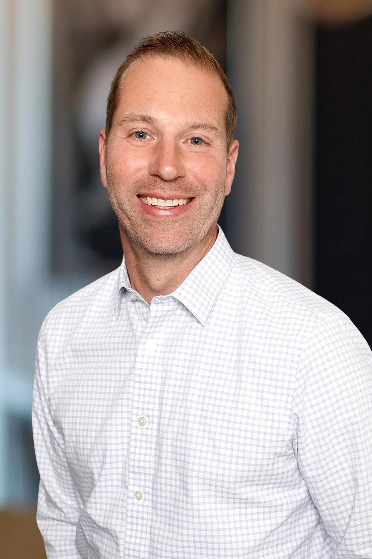 Headshot of a smiling man with short brown hair, wearing a light-colored checkered button-up shirt, standing indoors with a blurred office background.