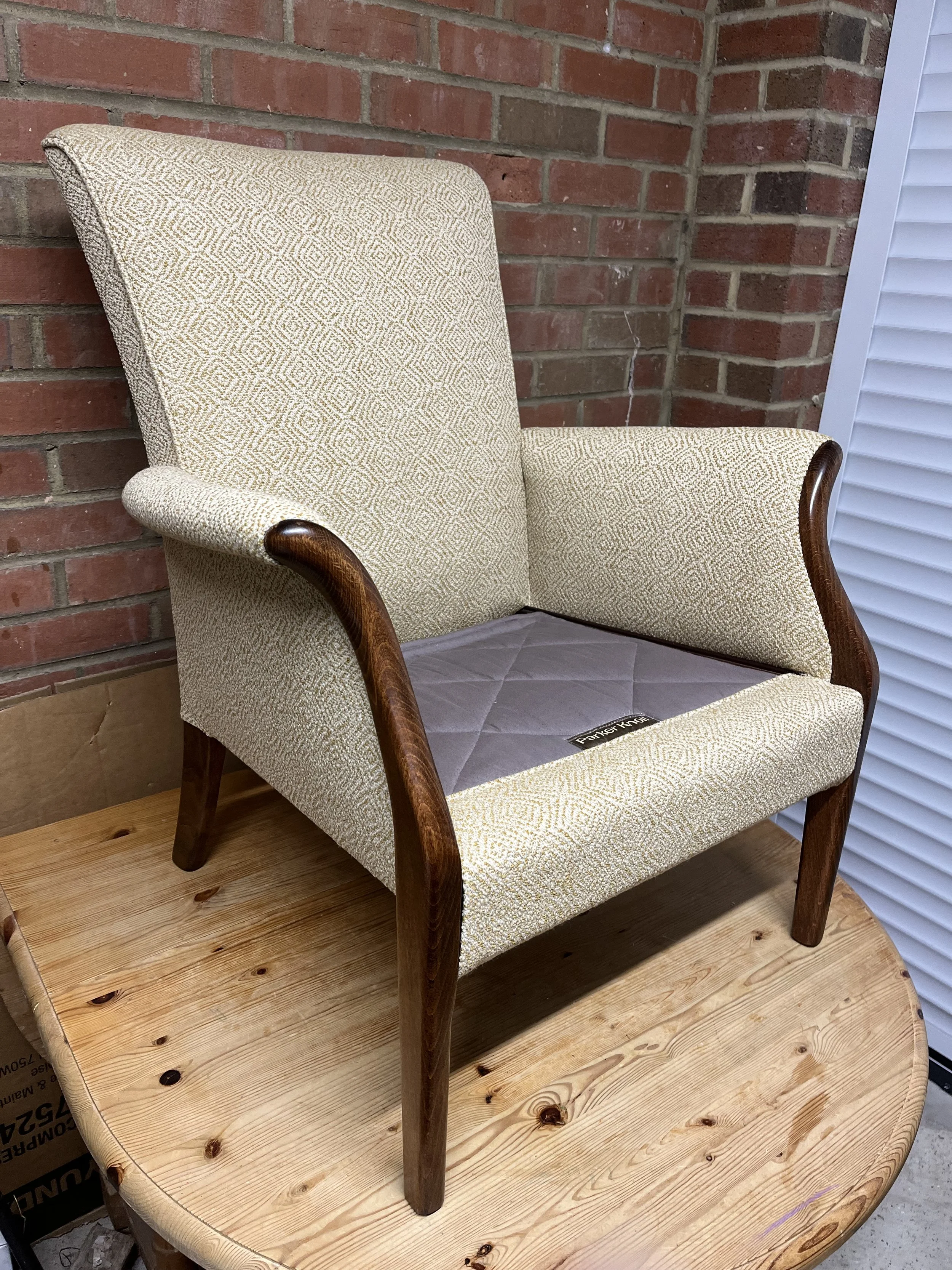 A vintage beige armchair with wooden armrests and legs sitting on a round wooden table with a brick wall background.