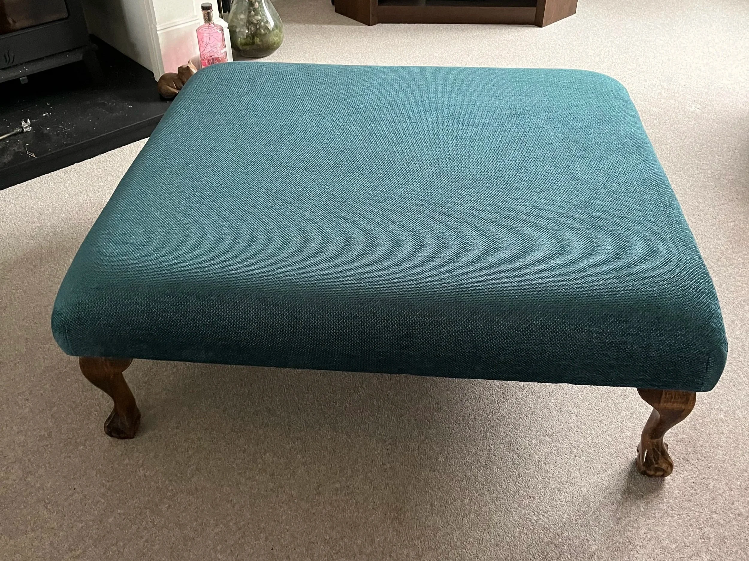 A wooden-legged ottoman with teal fabric upholstery on a beige carpeted room floor.