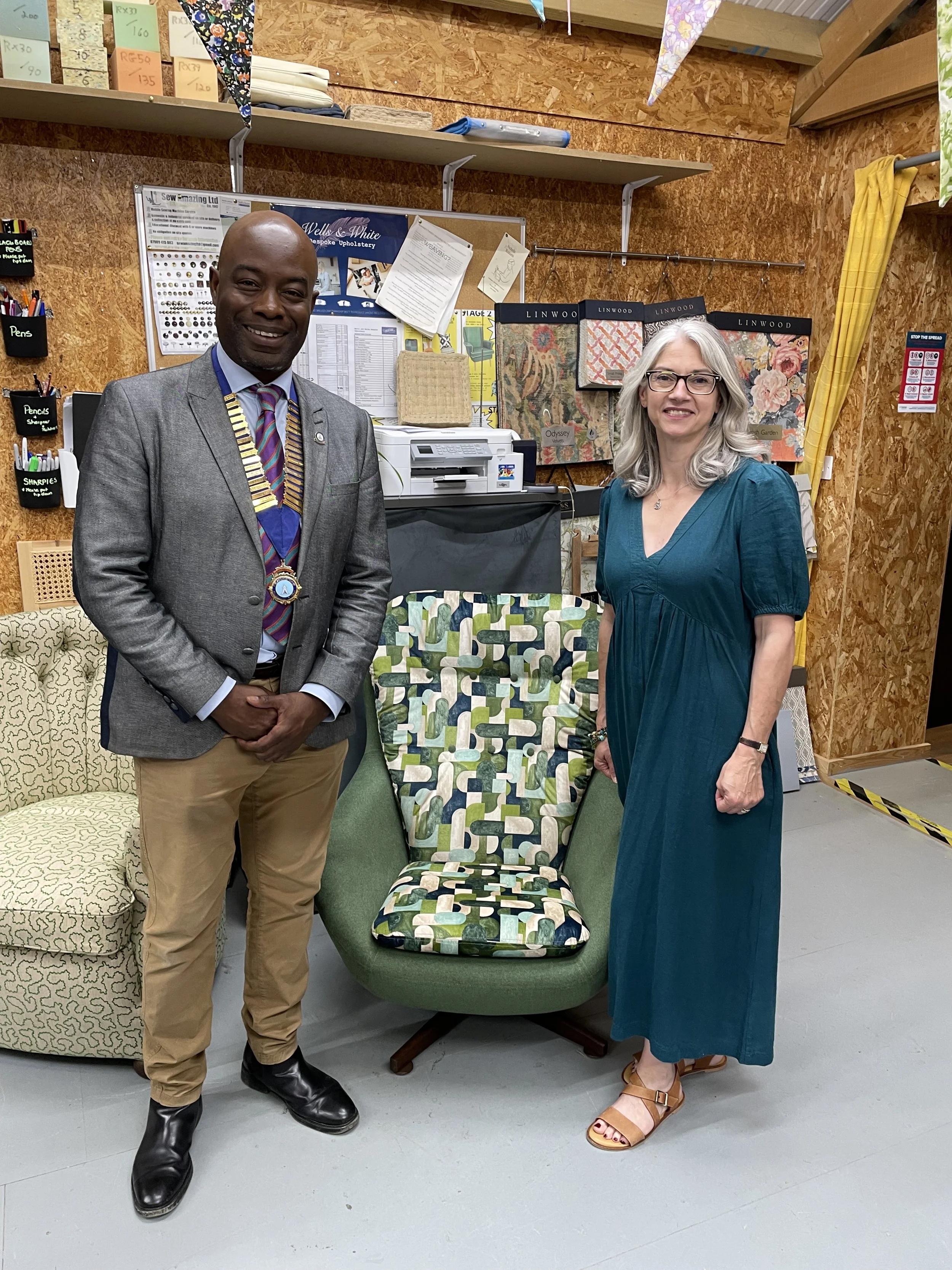 Two people, a man and a woman, standing inside a store or workshop with shelves of fabric and upholstery samples behind them. The man wears a gray suit, colorful tie, and a chain with a medallion. The woman wears glasses and a teal dress, with sandals. They are standing beside a green and patterned upholstered chair.