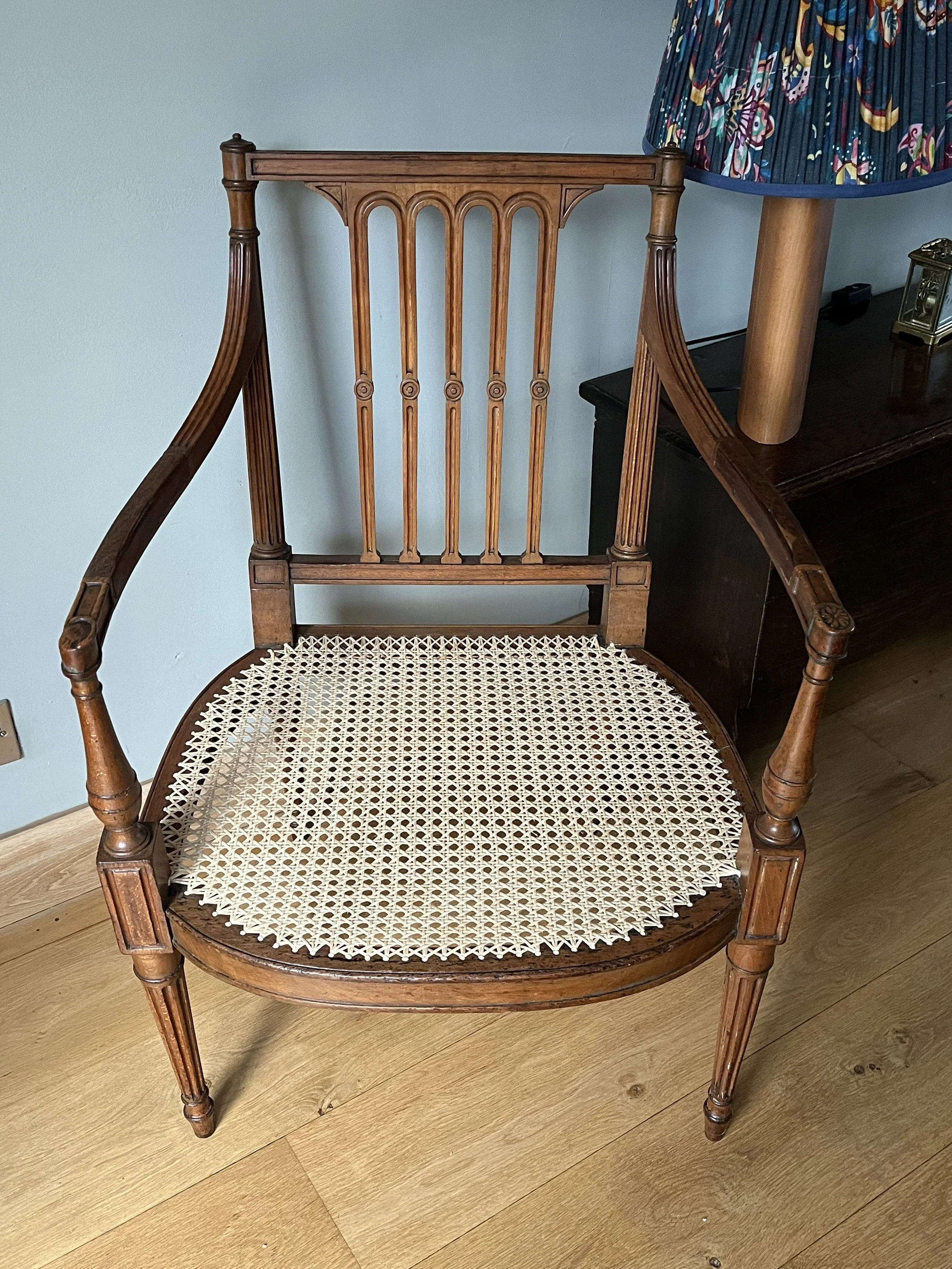 A wooden chair with a woven cane seat in a room with hardwood flooring. Part of a table with a colorful lamp and some small objects are visible on the right.