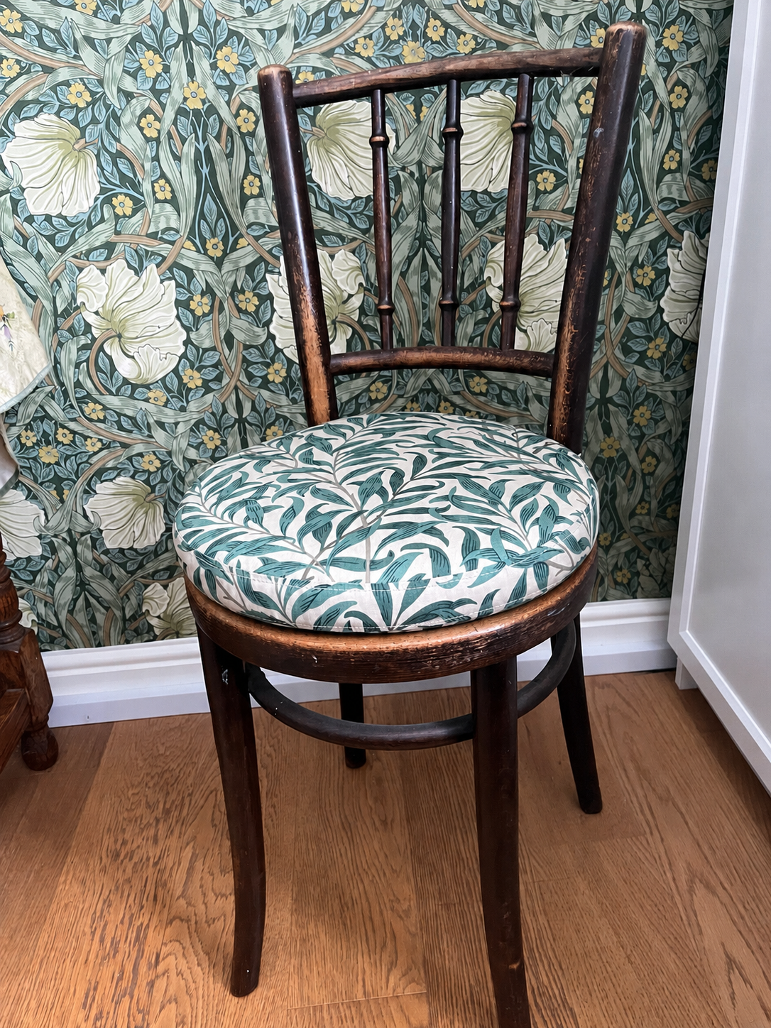 A vintage wooden chair with a round, fabric-covered cushion featuring a green and white leaf pattern, placed on a wooden floor against a floral wallpaper background with large white flowers and green leaves.
