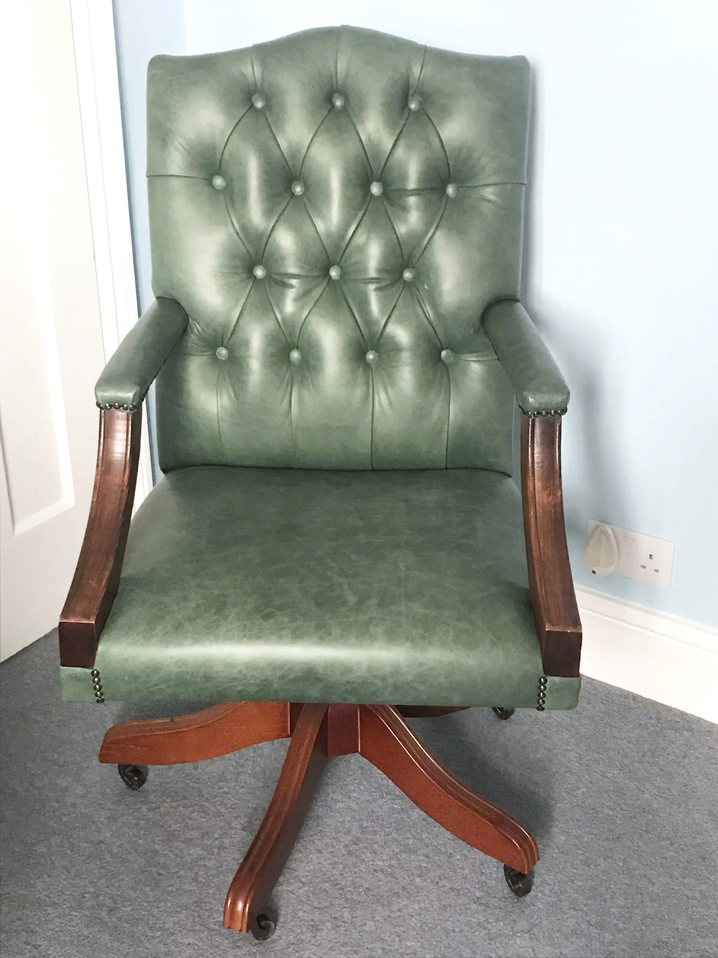 A green leather high-back armchair with wooden armrests and a wooden base on wheels, positioned on a carpeted floor against a light-colored wall.