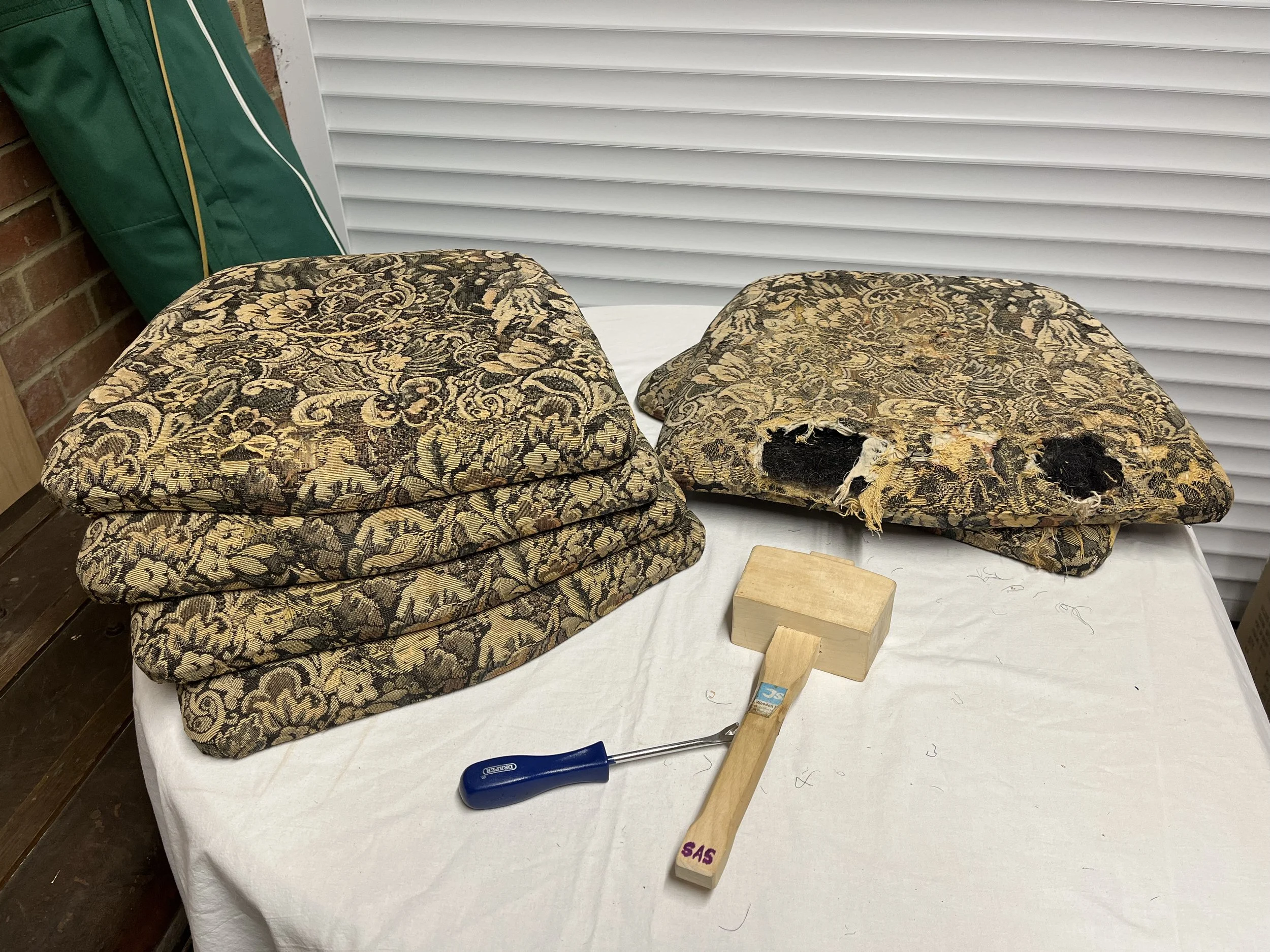 Two damaged floral-patterned upholstered chairs with torn fabric, placed on a white table. A hammer and a wooden block are on the table nearby.