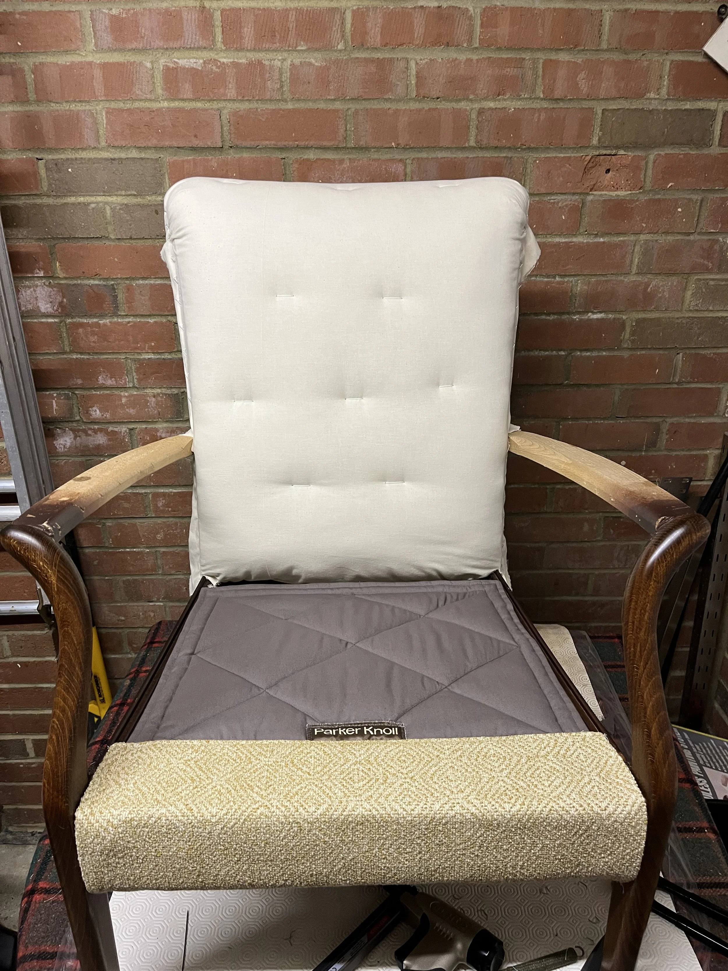 A wooden armchair with a white cushion and a yellow seat cushion, set against a brick wall. In front of the chair is a black mat with some tools including a cordless drill and a leveling tool.