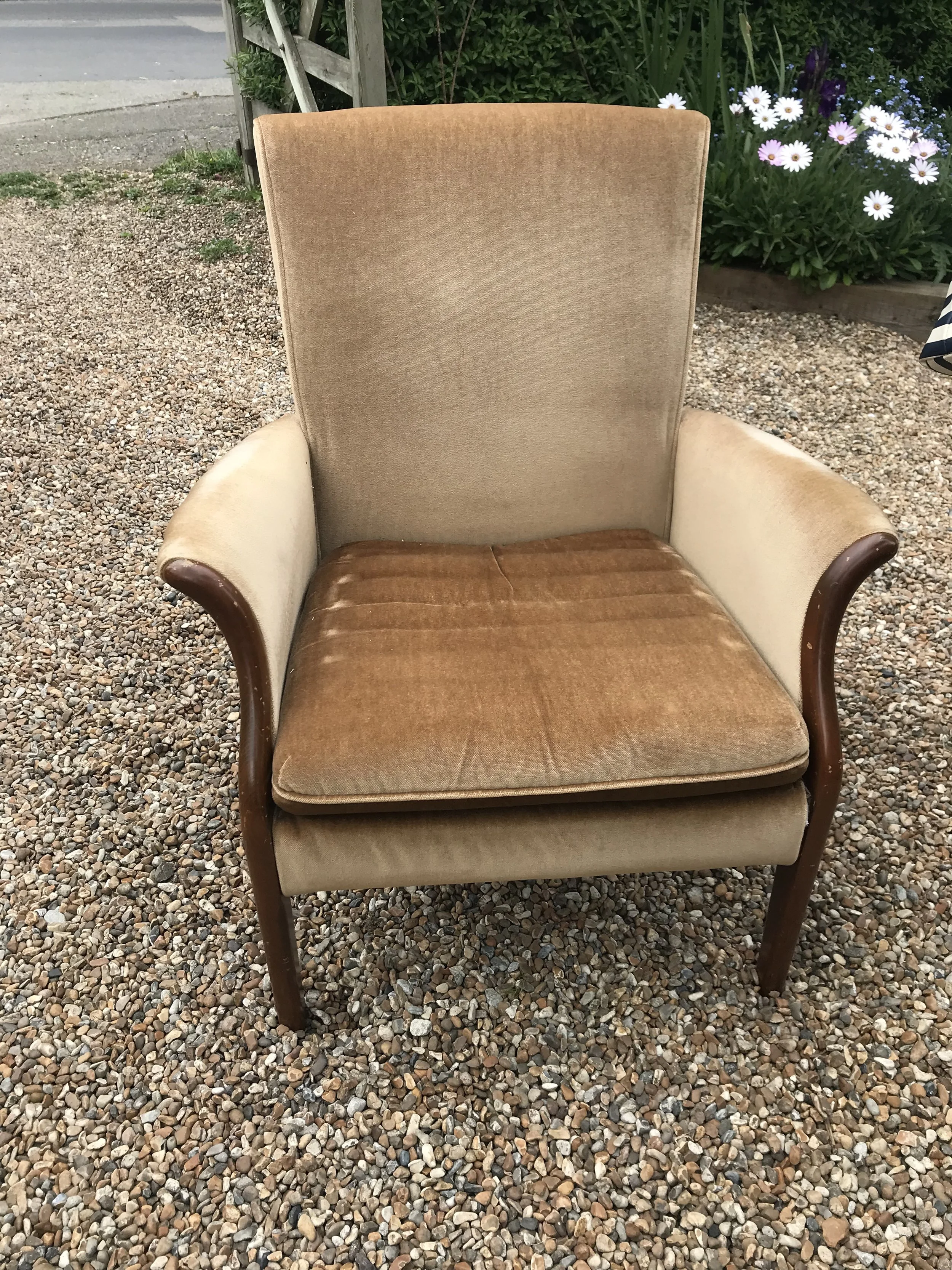A vintage armchair with beige upholstery and wooden armrests and legs, placed outdoors on gravel with plants and flowers in the background.
