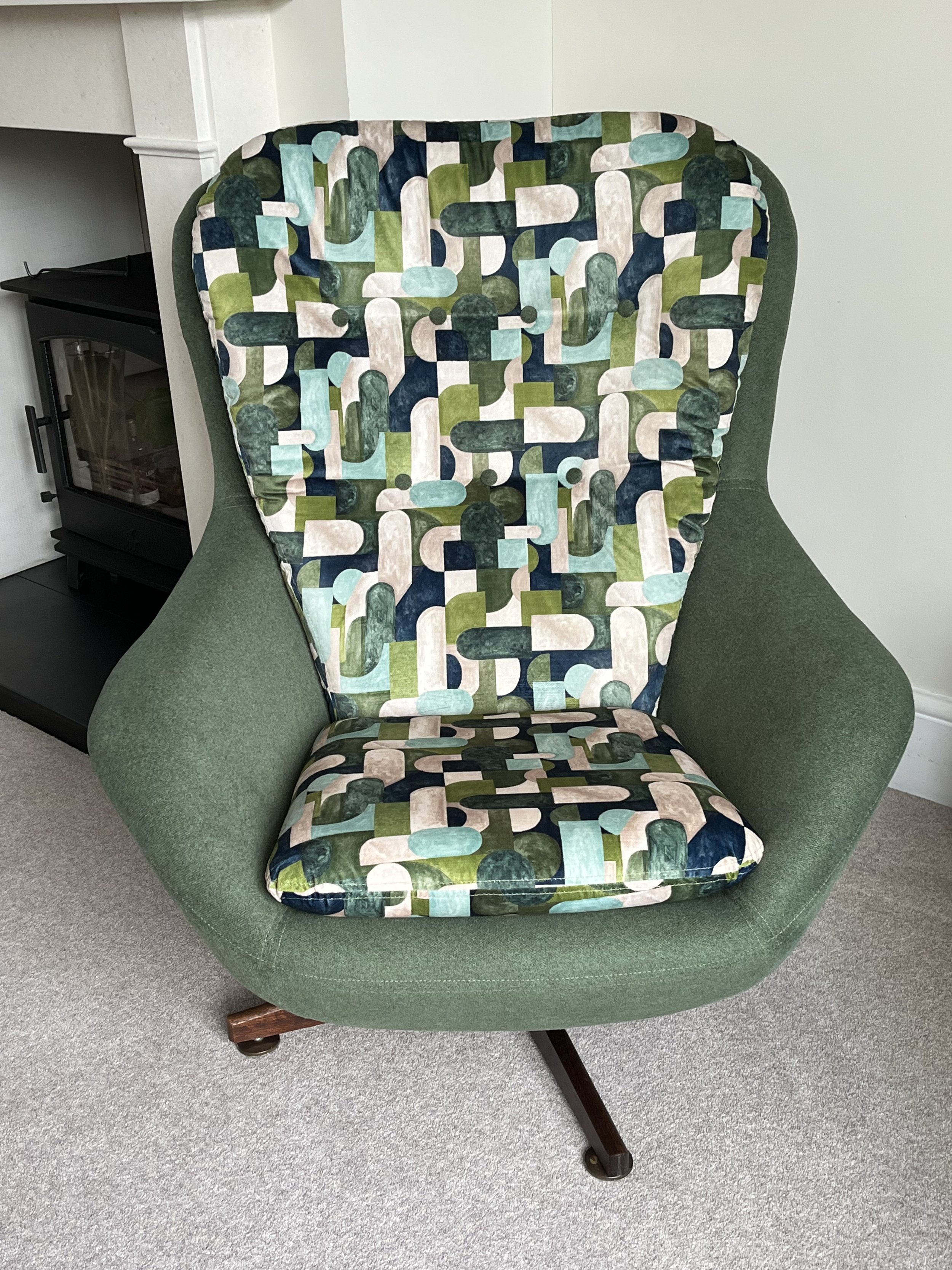 A green upholstered armchair with wooden legs, featuring a cushion and backrest with a geometric pattern of various shades of green, white, blue, and beige.