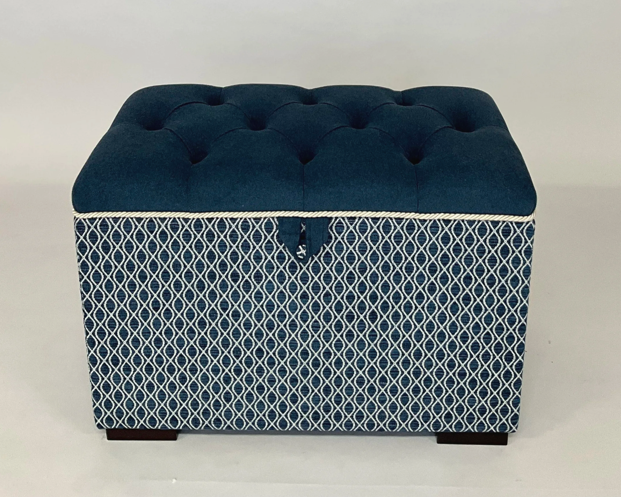 A square, blue upholstered ottoman with a diamond pattern fabric base, a cushioned top with tufted buttons, and dark wooden feet.