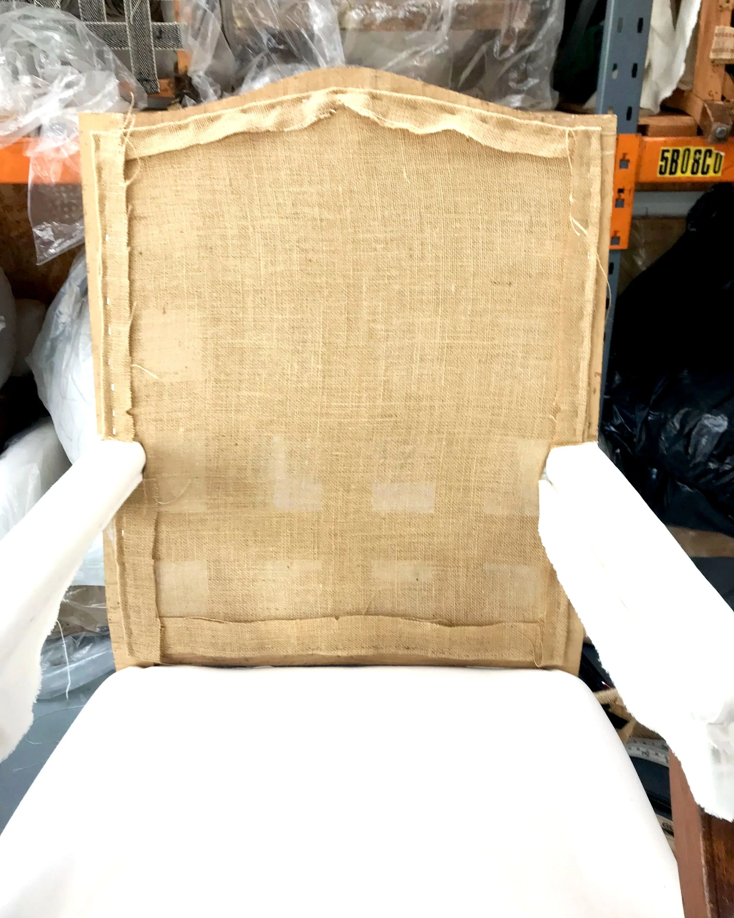 Back of an upholstered piece, showing fabric and edge detailing, in a workspace surrounded by storage and supplies.