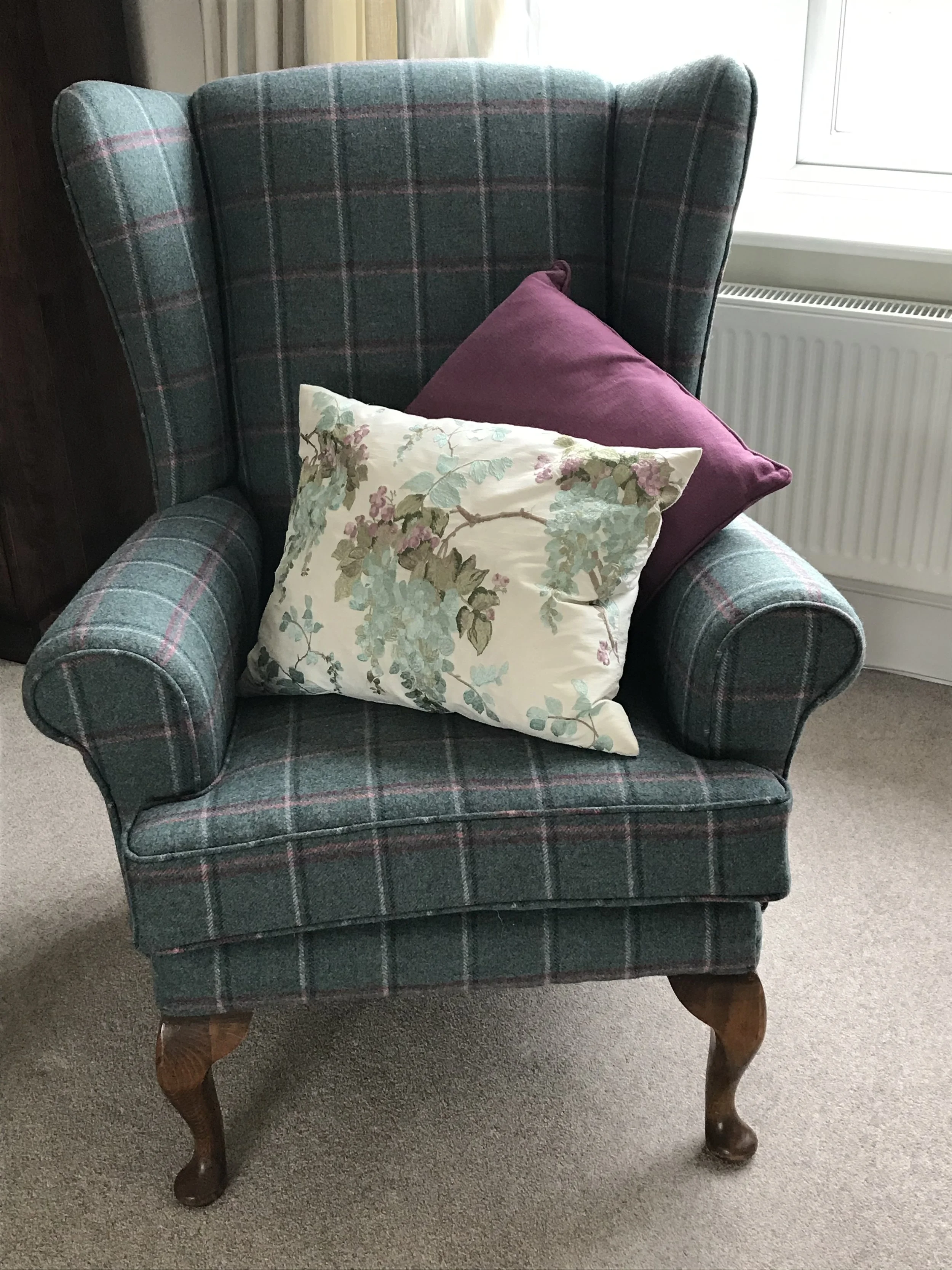 A vintage-style armchair with wooden legs and a plaid fabric pattern in shades of green, blue, and pink. Two decorative pillows, one dark purple and one floral with pastel colors, are placed on it. The chair is positioned on a beige carpet near a window with a white radiator underneath.