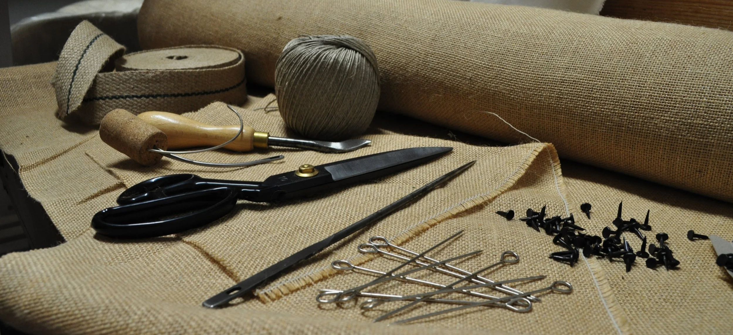 A collection of sewing tools and materials on a burlap surface, including scissors, a seam ripper, a ball of gray thread, gray fabric, and pins.