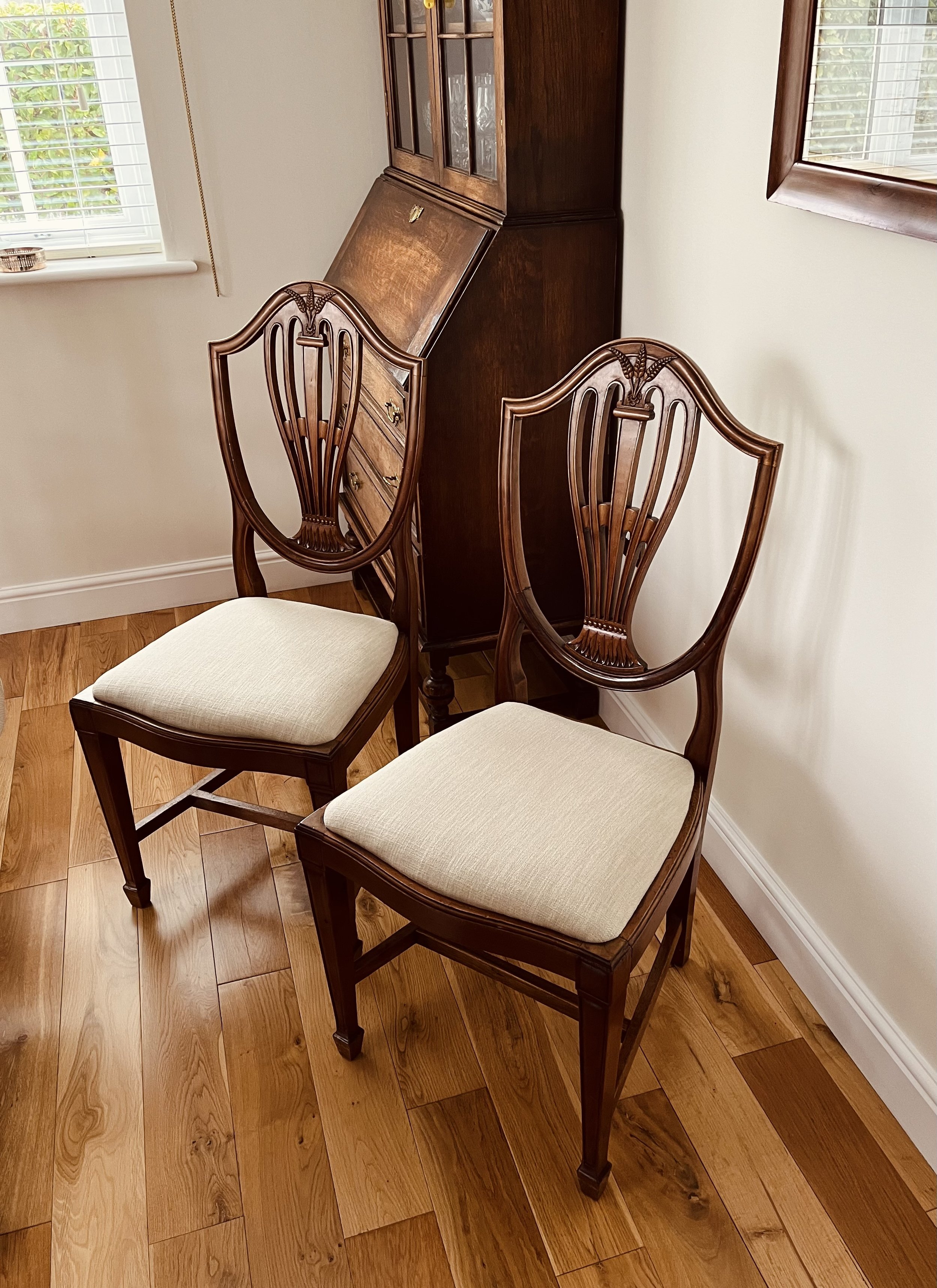 Two wooden chairs with beige cushioned seats in a room with hardwood flooring, a window with white blinds, and a wooden china cabinet against the wall.