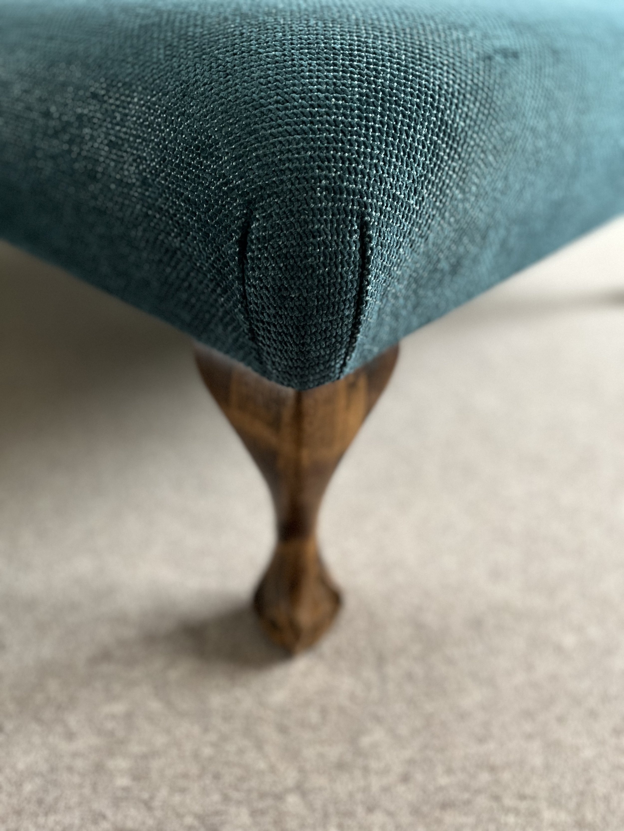 Close-up of the corner of a teal-colored fabric-covered piece of furniture, with a wooden leg visible underneath, on a light-colored surface.