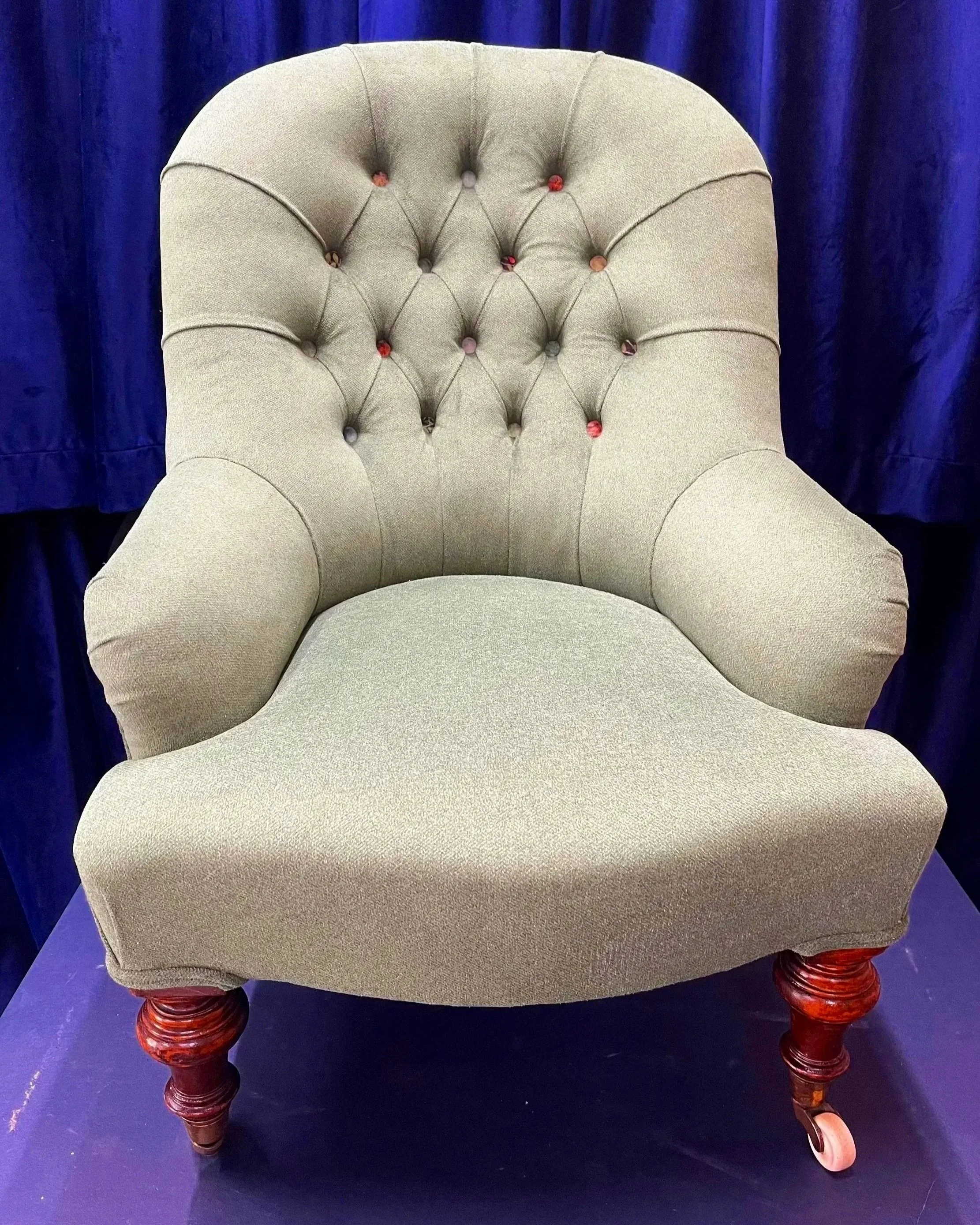 A beige upholstered armchair with button tufting on the backrest, set against a dark blue curtain background. The chair has curved armrests and four carved wooden legs with a dark finish.