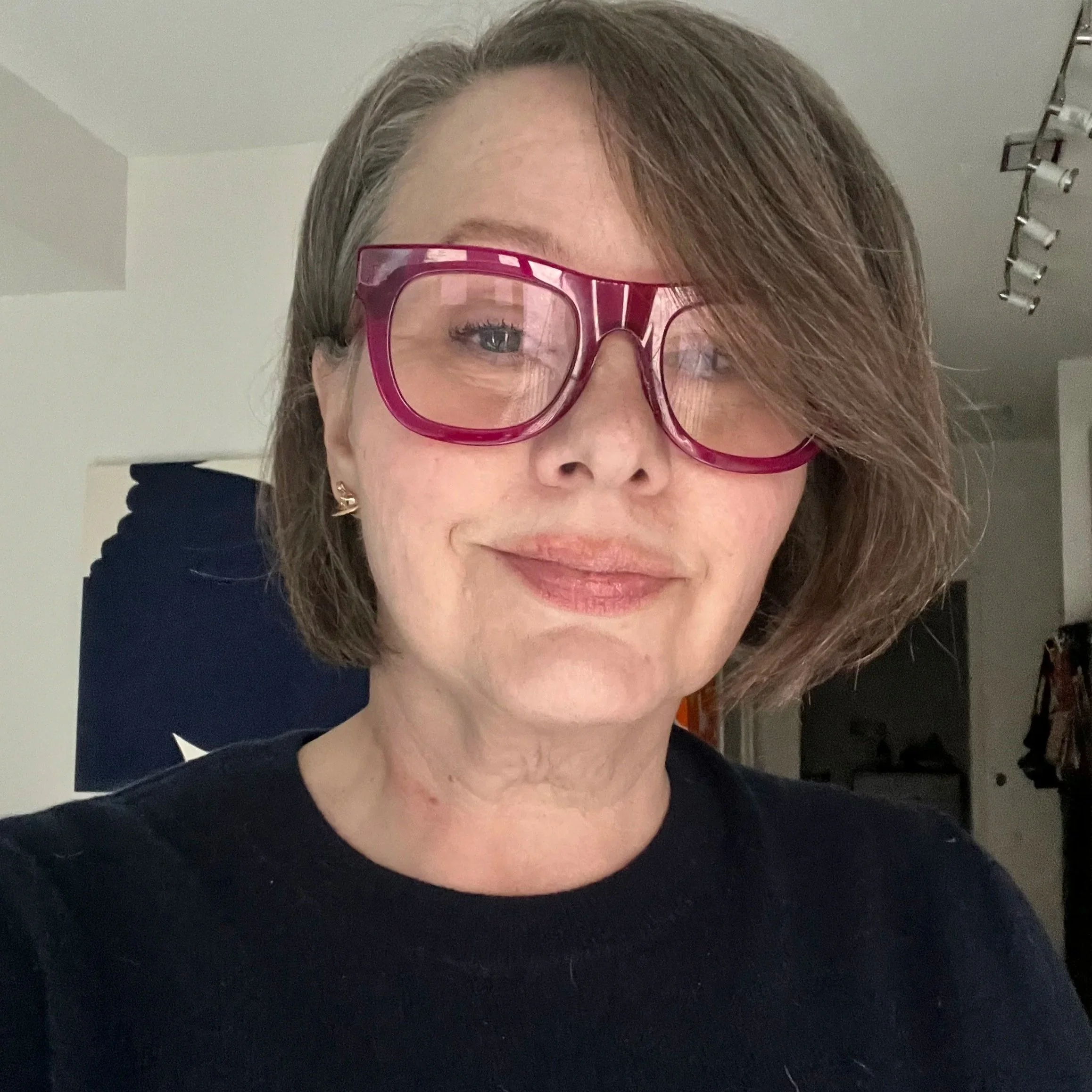 Close-up of a woman wearing glasses with a reflection of a window, with brown hair, earrings, and a black top, indoors with gray walls and ceiling lights.