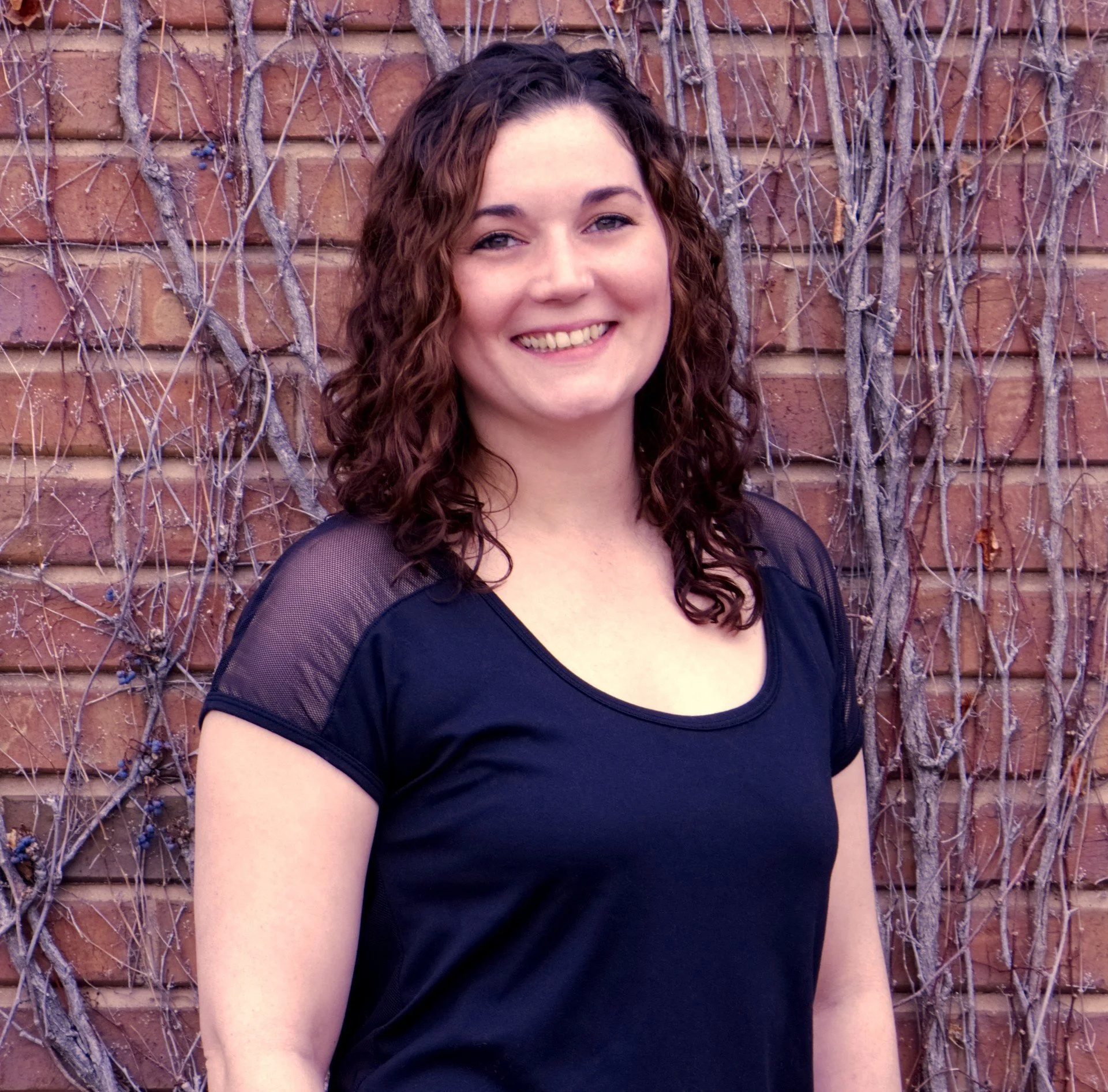A woman with curly brown hair and fair skin smiling at the camera, wearing a black top with sheer sleeves, standing in front of a brick wall covered with dry, leafless vines.