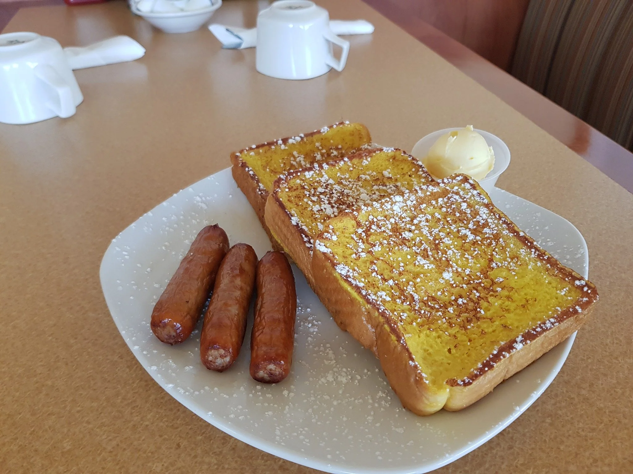 A plate with French toast topped with powdered sugar, served with a side of mashed potatoes, along with sausage links. In the background, there are cups and bowls on a wooden table.