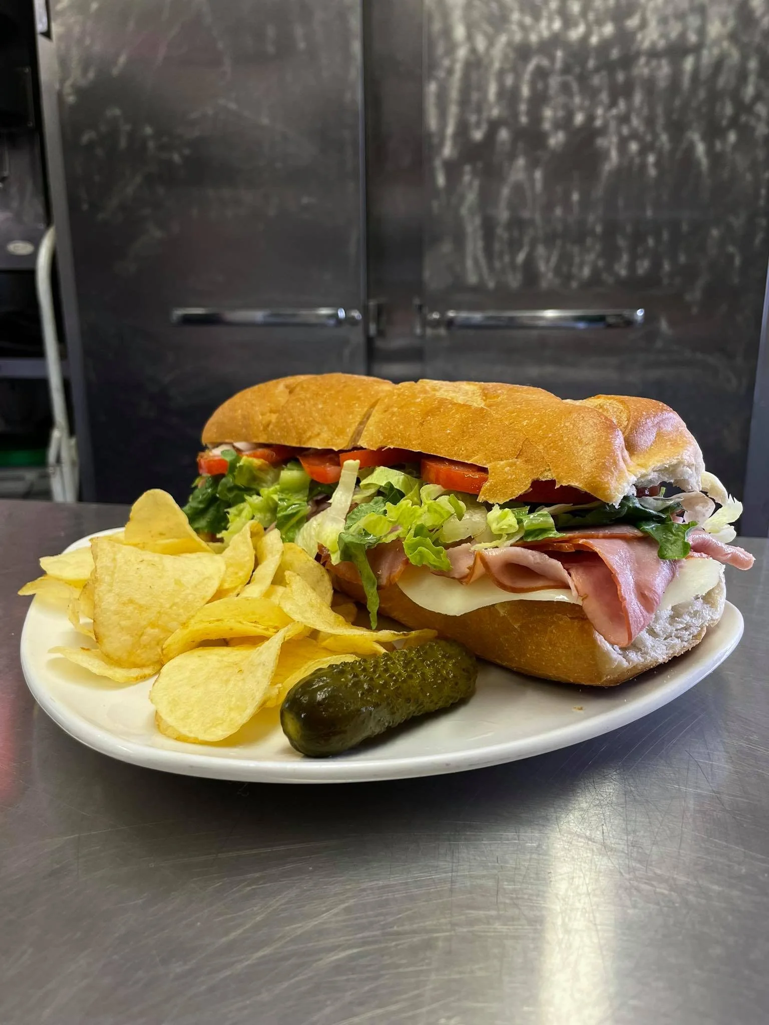 A plate with a submarine sandwich containing lettuce, tomato, ham, cheese, and a hoagie bun, served with potato chips and a pickle, on a stainless steel surface.