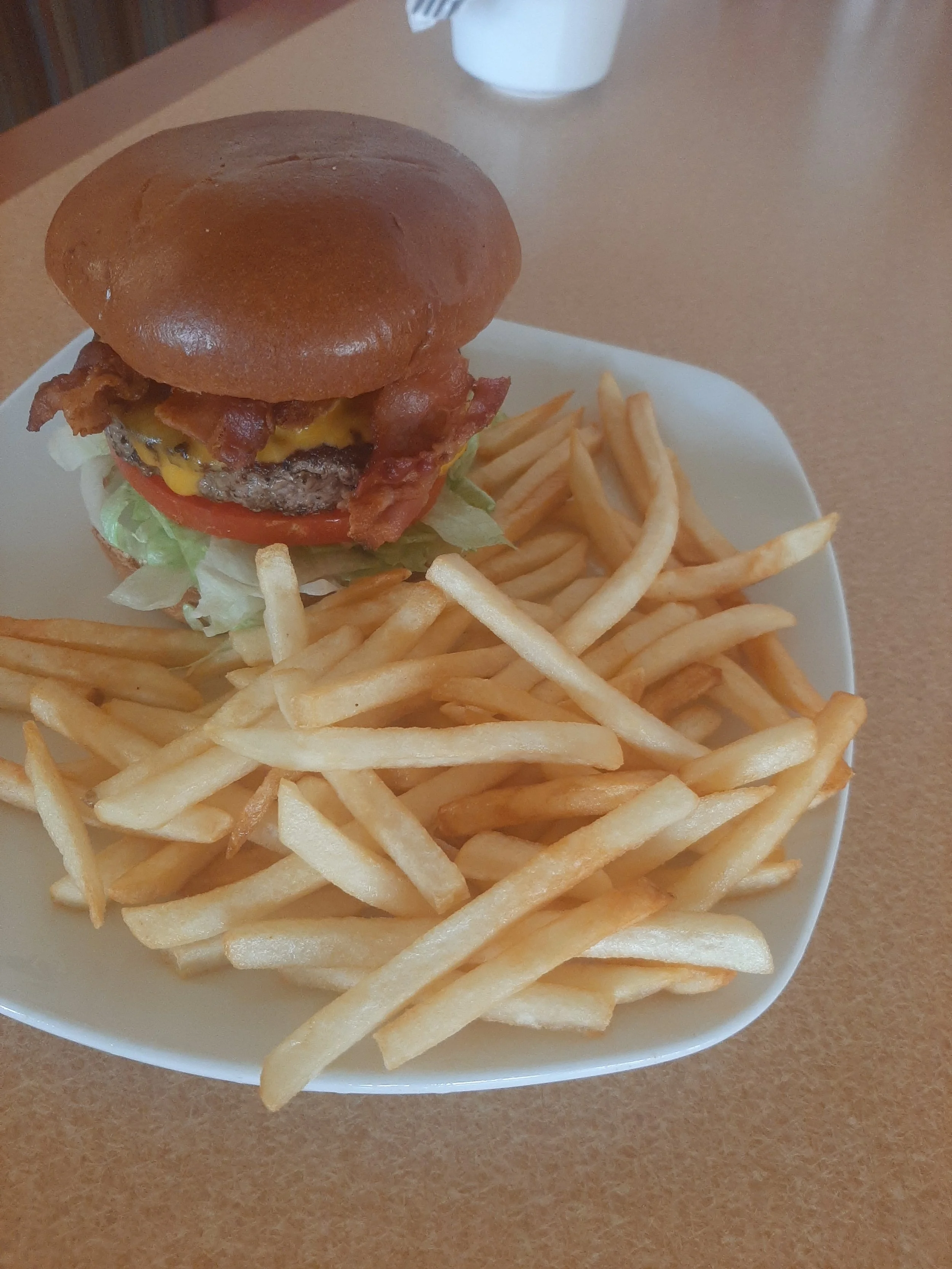 Plate of French fries with a cheeseburger, lettuce, tomato, bacon, and a bun, on a brown table.