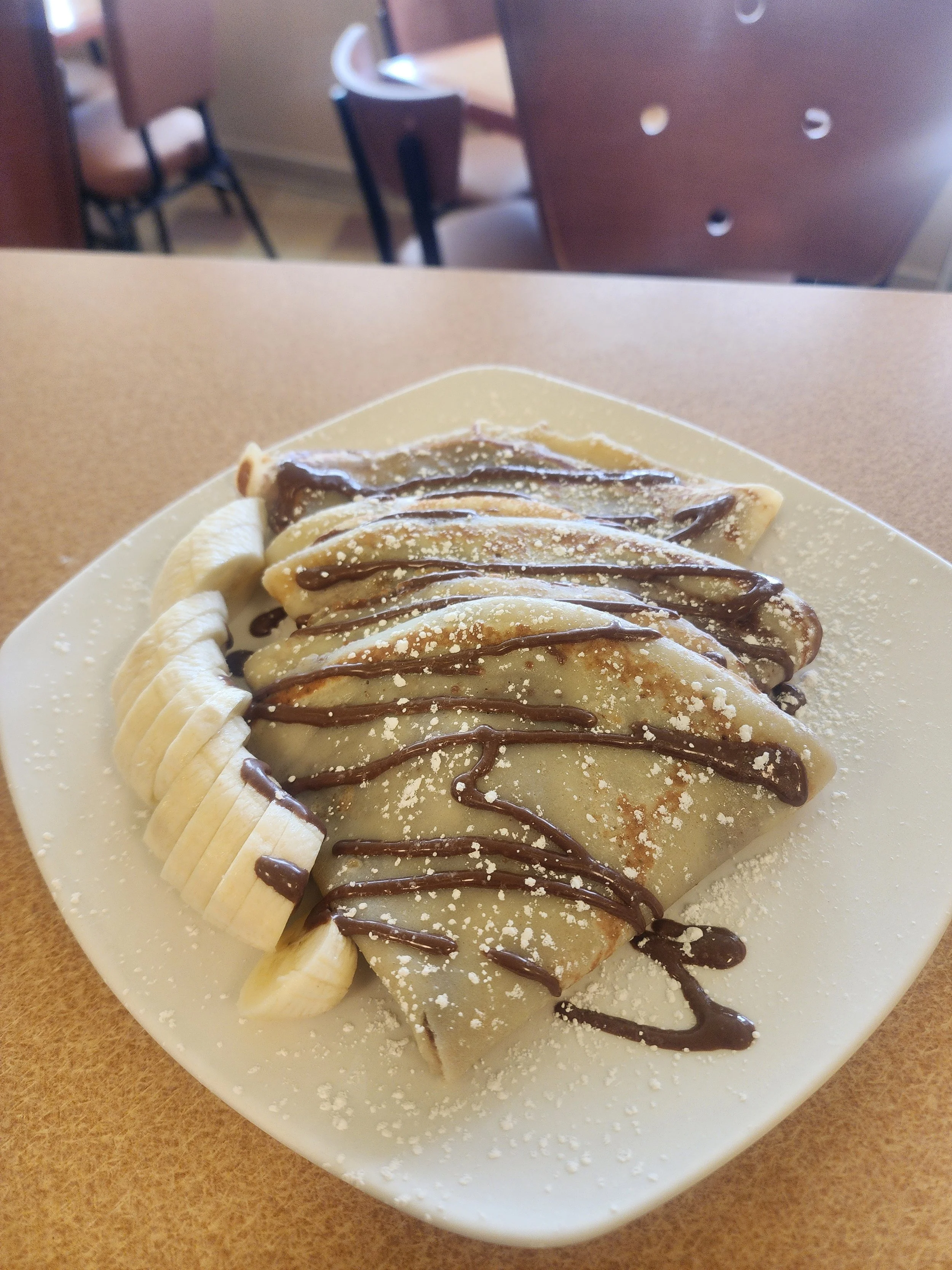 A dessert crepe drizzled with chocolate sauce and topped with powdered sugar, served with sliced bananas on a white plate.