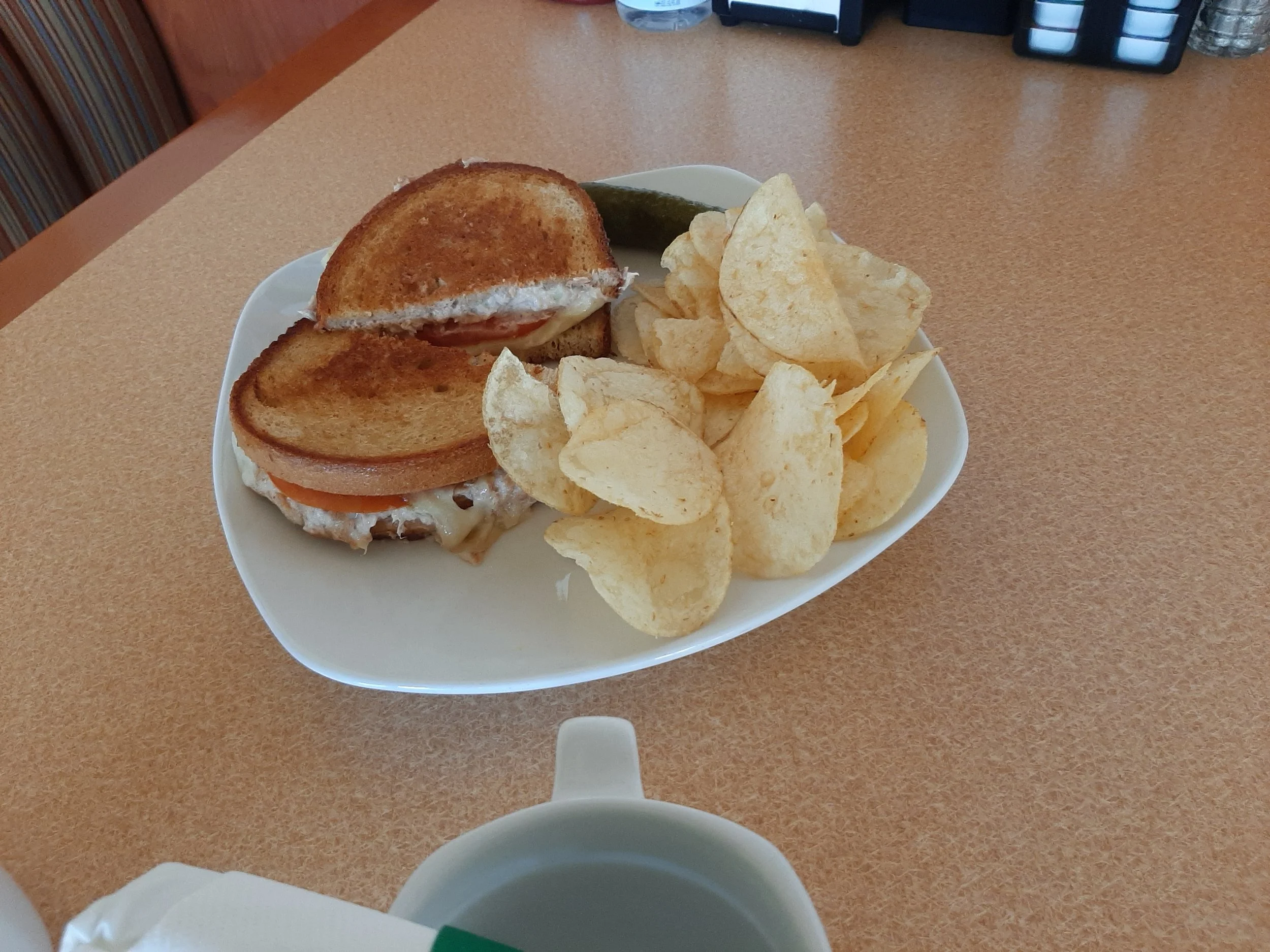A plate with a partially eaten club sandwich containing tomato, cheese, and turkey, served with potato chips and a pickle on a diner table.