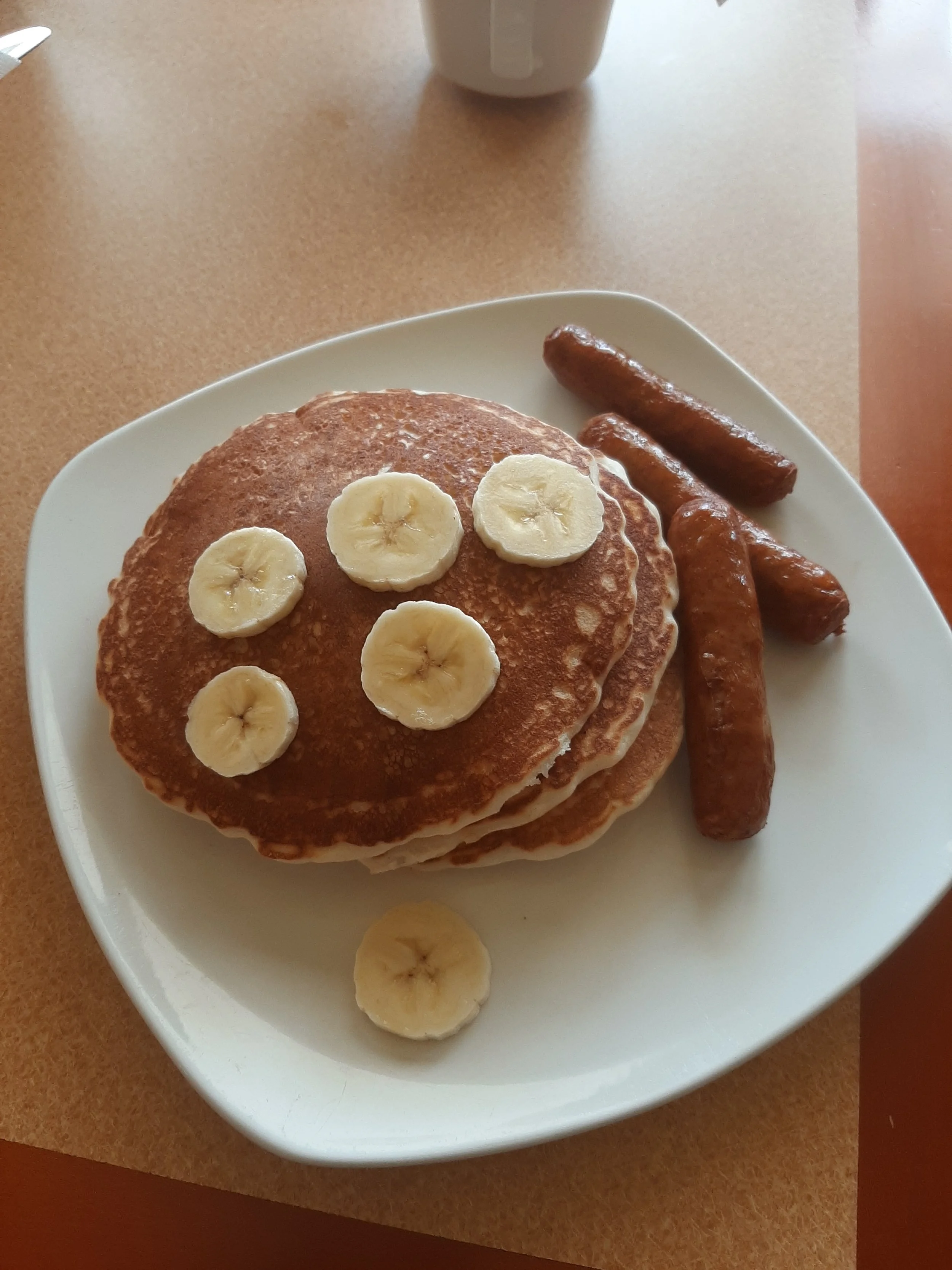 Plate with three pancakes topped with banana slices and three sausage links on the side.