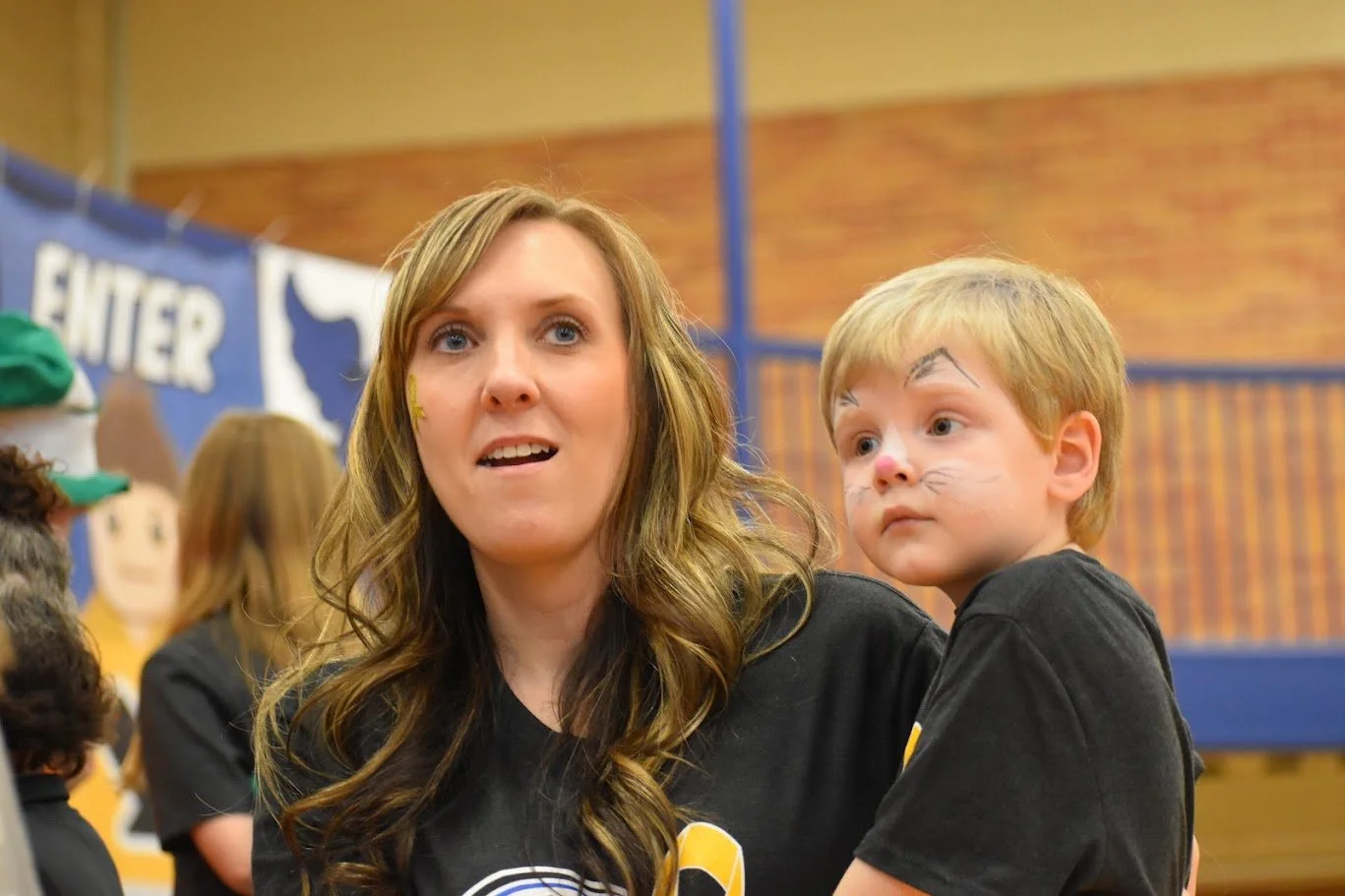 A woman with long wavy brown hair and a yellow star sticker on her cheek, and a young boy with face paint of a cat, are together indoors at a school or event.