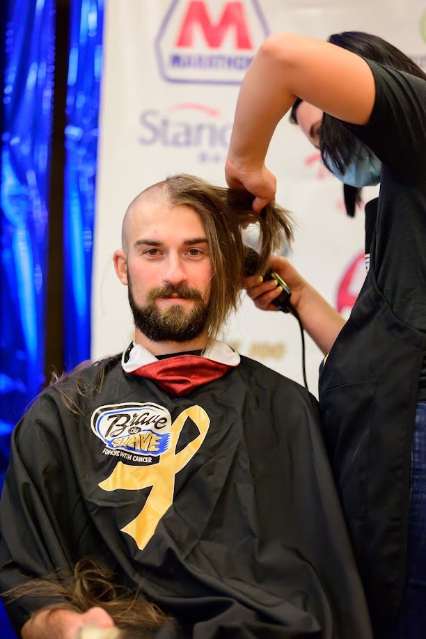 A man with long hair is getting his hair cut at a charity event supporting cancer awareness, with a woman wearing a face mask styling his hair.
