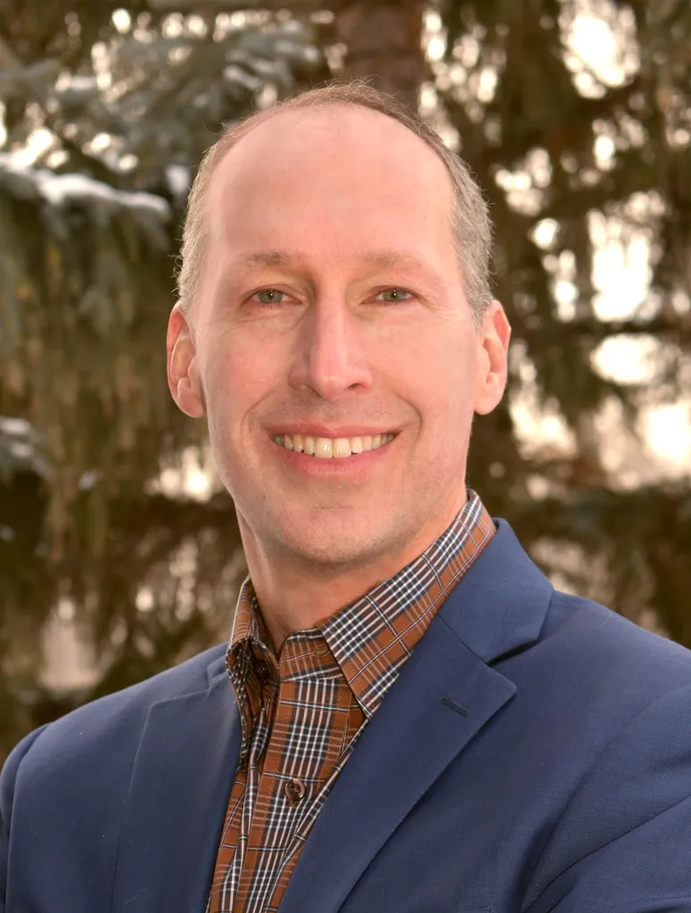 A man in a blue blazer and plaid shirt smiling outdoors with trees in the background.