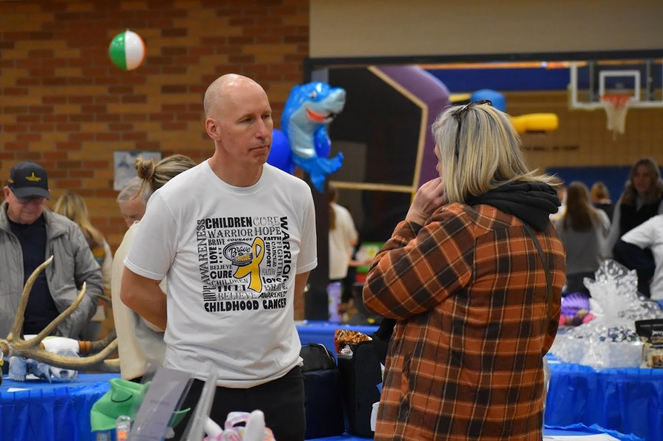A man and a woman are having a conversation at a event with blue tables and decorations. The man is wearing a white t-shirt with yellow ribbon and words related to childhood cancer awareness. The woman is in a brown and black plaid hoodie. There are other people, balloons, and a basketball hoop in the background.